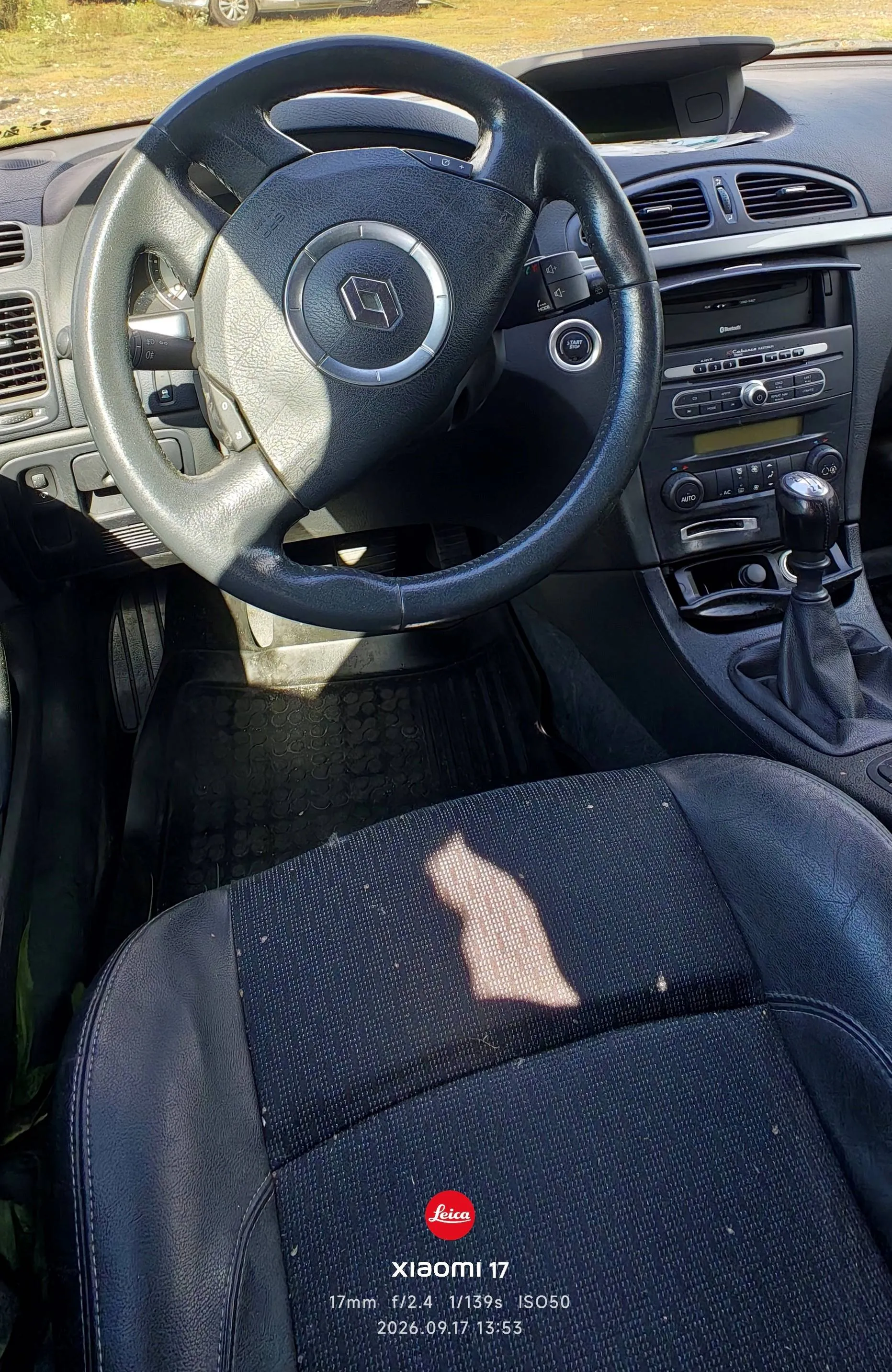 Widok z wnętrza samochodu z perspektywy kierowcy. W górnej części zdjęcia widoczna jest czarna kierownica z logo marki Renault w centrum. Po prawej stronie znajduje się konsola środkowa z radiem, przyciskami sterowania i lewarkiem zmiany biegów. W dolnej części obrazu widoczny jest fragment czarnego, skórzanego fotela kierowcy z jasną plamą na oparciu. Na dole zdjęcia znajduje się biały napis: 'Leica xiaomi 17 17mm f/2.4 1/139s ISO50 2026.09.17 13:53'.