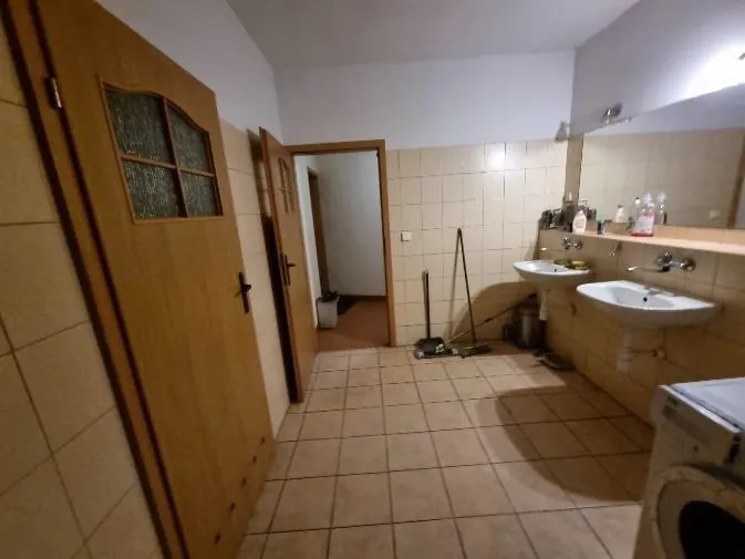 Wnętrze łazienki z beżowymi płytkami na ścianach i podłodze. Po prawej stronie znajdują się dwa umywalki z lustrem powyżej, na którym stoją butelki i przedmioty toaletowe. Po lewej stronie widoczne są drewniane drzwi z matowymi szybkami oraz otwarte drzwi prowadzące do innego pomieszczenia. W głębi pokoju stoi mop z wiadrem, a w prawym dolnym rogu znajduje się pralka. Ściany są wyłożone jasnymi płytkami, a sufit jest biały.