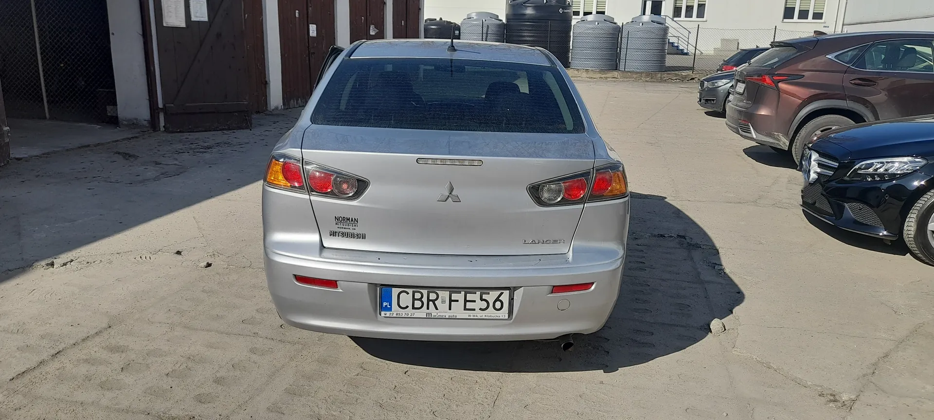 Tył srebrnego samochodu Mitsubishi Lancer z tablicą rejestracyjną CBR FE56 (PL). Na bagażniku widoczne napisy 'NORMAN MITSUBISHI' oraz logo marki. W tle parking z innymi pojazdami (m.in. brązowy SUV, czarny samochód), przemysłowe budynki z bramami, zbiornikami i ogrodzeniem. Asfaltowa powierzchnia parkingu z widocznymi pęknięciami.