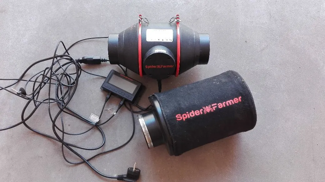 Zestaw urządzeń marki Spider Farmer ułożony na szarym podłożu. W górnej części znajduje się czarny wentylator kanałowy z białym paskiem z instrukcjami i czerwonym logo 'Spider Farmer'. Poniżej widoczny jest cyfrowy sterownik z ekranem i przyciskami oraz czarny filtr węglowy w kształcie cylindra z czerwonym napisem 'Spider Farmer'. Całość połączona jest czarnymi przewodami elektrycznymi z wtyczką sieciową na końcu.