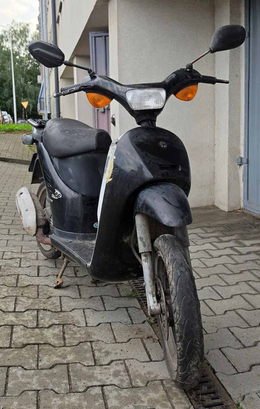 Czarny skuter zaparkowany na kostce brukowej przed jasną ścianą budynku. Pojazd jest w widocznym stanie zużycia, z zabrudzeniami na nadwoziu i kołach. Widoczny jest reflektor, dwa pomarańczowe kierunkowskazy oraz lusterka boczne.