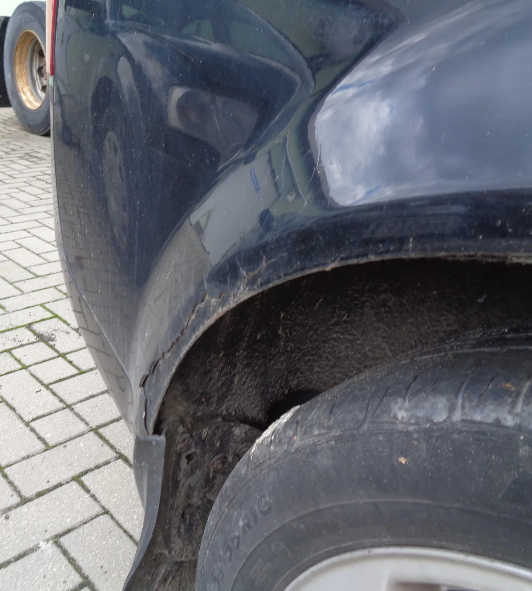 Zbliżenie na uszkodzony błotnik czarnego samochodu nad przednim kołem. Widoczne jest pęknięcie i ubytek lakieru wzdłuż krawędzi nadkola, gdzie plastikowy element łączy się z karoserią. W tle widoczna jest kostka brukowa.