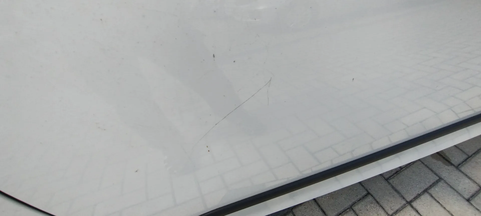 Zbliżenie na białą, błyszczącą powierzchnię karoserii samochodu. Na lakierze widoczna jest cienka, ukośna rysa. W powierzchni odbija się wzór kostki brukowej z chodnika.