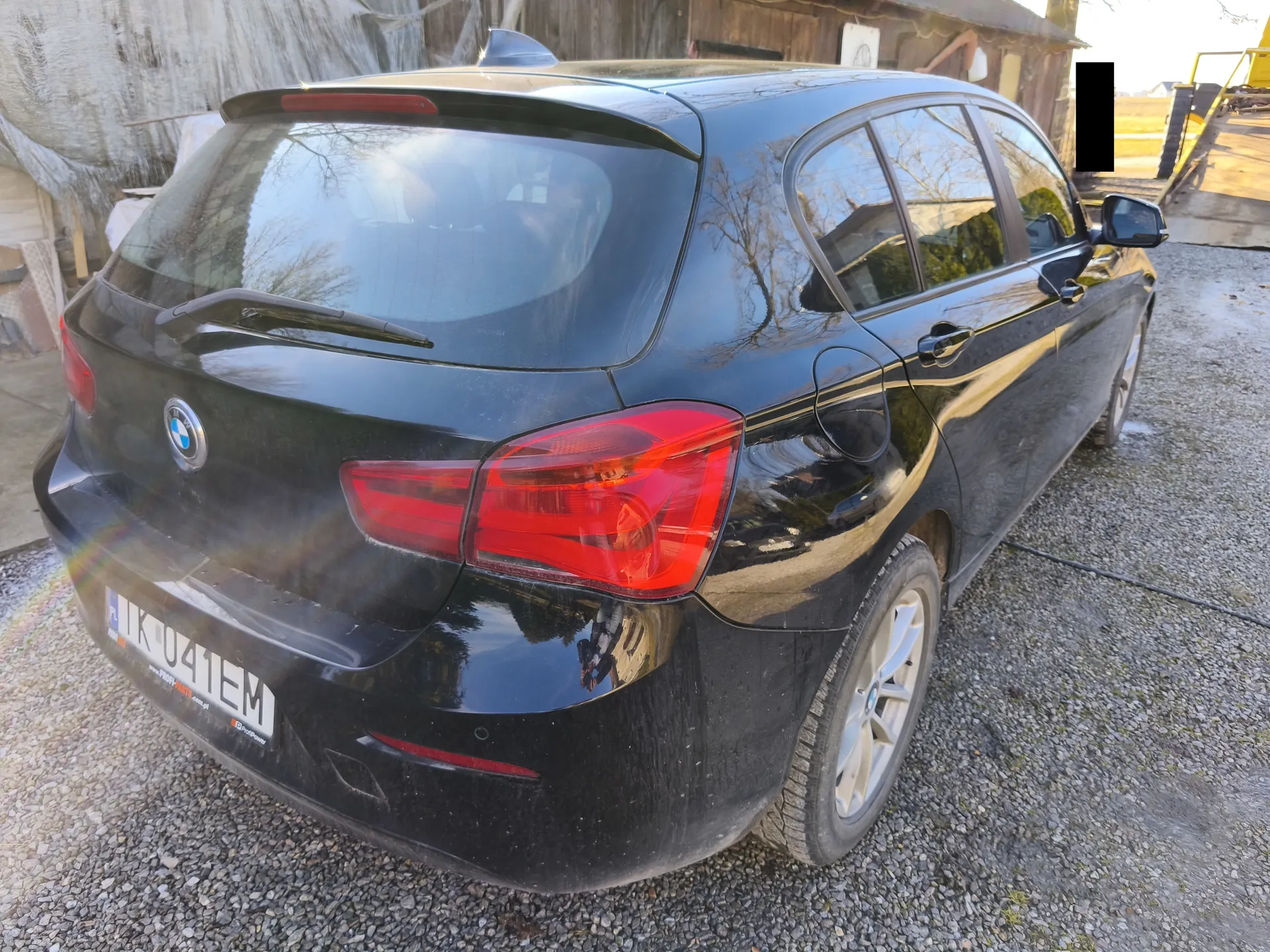 Czarny samochód osobowy marki BMW, widok z tyłu i boku. Widoczne czerwone światła tylne, logo BMW na bagażniku oraz tablica rejestracyjna z numerem IK 041EM. Samochód zaparkowany na żwirowym podłożu, w tle drewniana budowla i fragment żółtej konstrukcji. Na szybie tylnej odbijają się drzewa i otoczenie.