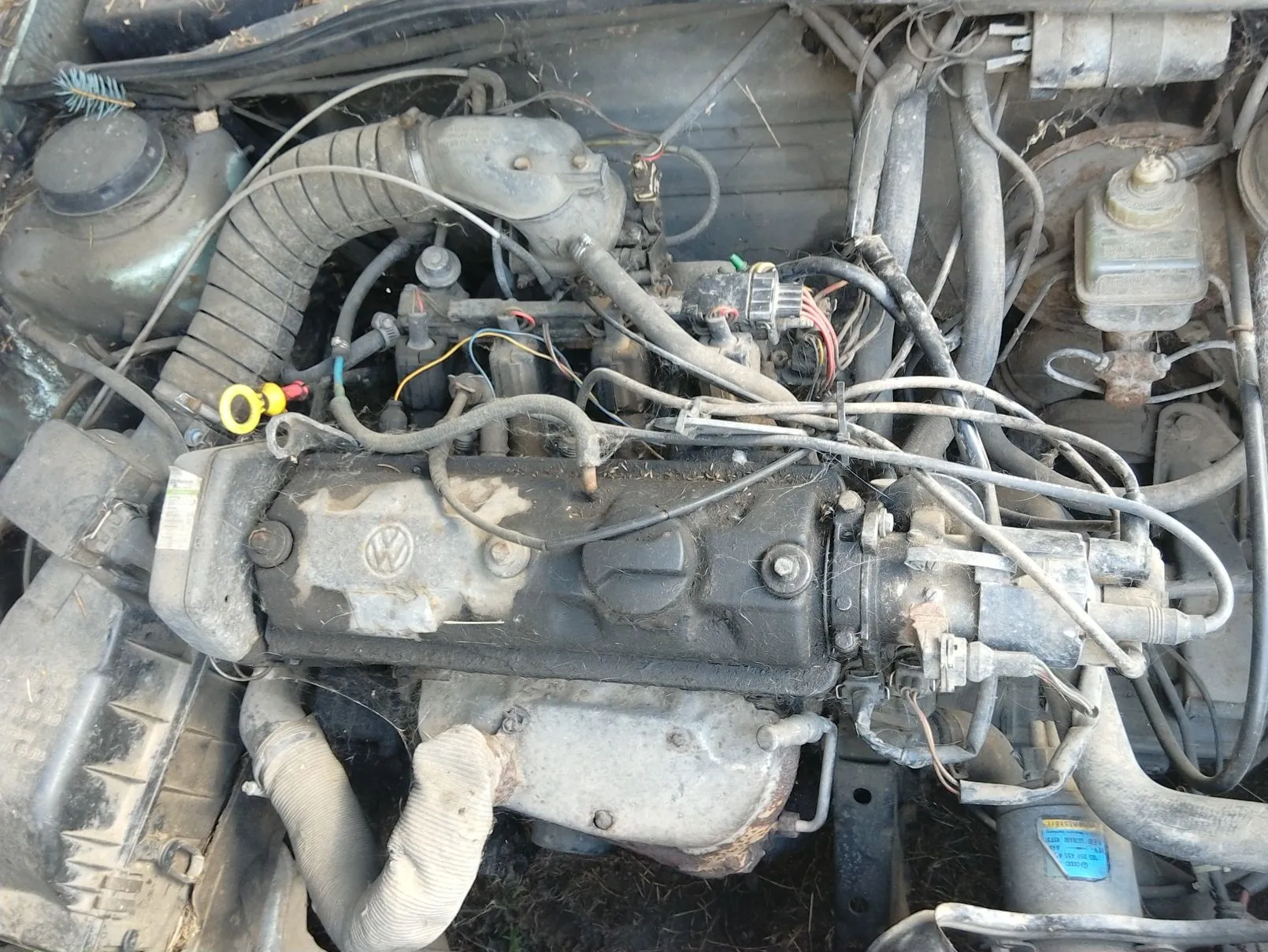 Widok z góry na zakurzony silnik samochodu marki Volkswagen, z widocznym logo VW na pokrywie zaworów. W komorze silnika znajdują się liczne przewody, rury dolotowe, wiązki elektryczne oraz zbiorniczki z płynami.