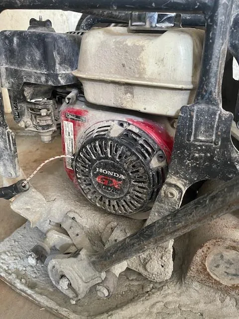 Zbliżenie na silnik marki Honda GX o czerwonym kolorze z białym zbiornikiem paliwa. Na silniku widoczny jest napis 'HONDA GX' oraz elementy mechaniczne pokryte kurzem i brudem. W tle częściowo widoczne są metalowe elementy konstrukcyjne i betonowa powierzchnia.