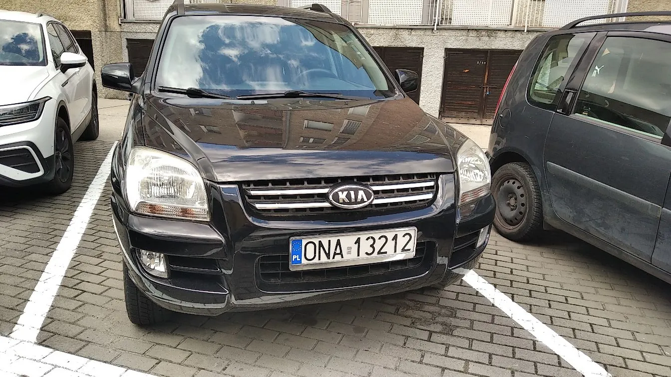 Czarny samochód marki Kia zaparkowany na kostce brukowej między białym a ciemnoszarym samochodem. Na przodzie pojazdu widnieje logo KIA oraz polska tablica rejestracyjna o numerze ONA 13212.