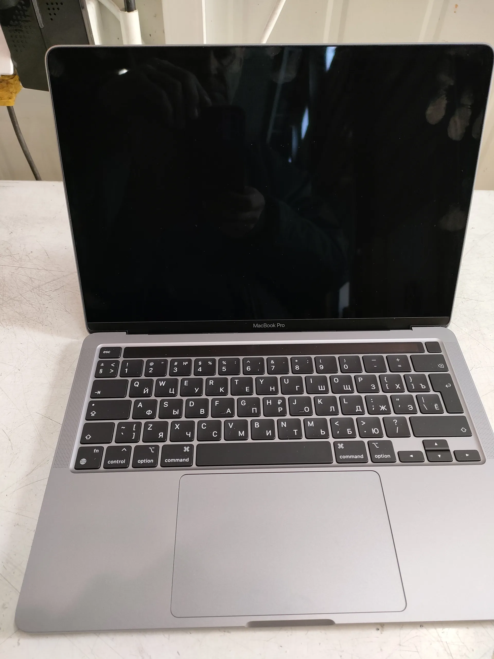 Otwarty laptop MacBook Pro z czarnym ekranem, na którym widoczne jest odbicie osoby trzymającej telefon. Klawiatura z czarnymi klawiszami zawiera znaki łacińskie i cyryliczne. Poniżej ekranu widnieje napis 'MacBook Pro'. Laptop znajduje się na białej, zadrapanej powierzchni. W tle widoczne są kable i biała konstrukcja.