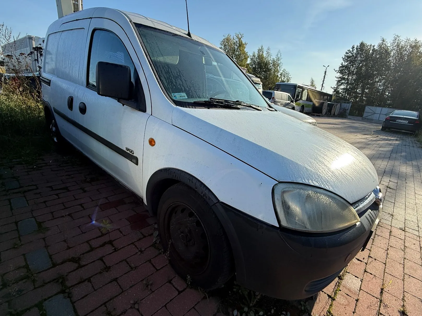 Biały samochód dostawczy marki Renault Kangoo zaparkowany na kostce brukowej. Pojazd jest widoczny z przodu i z boku, na karoserii widoczna jest rosa. W tle znajdują się inne samochody, autobus oraz drzewa pod jasnym, niebieskim niebem.