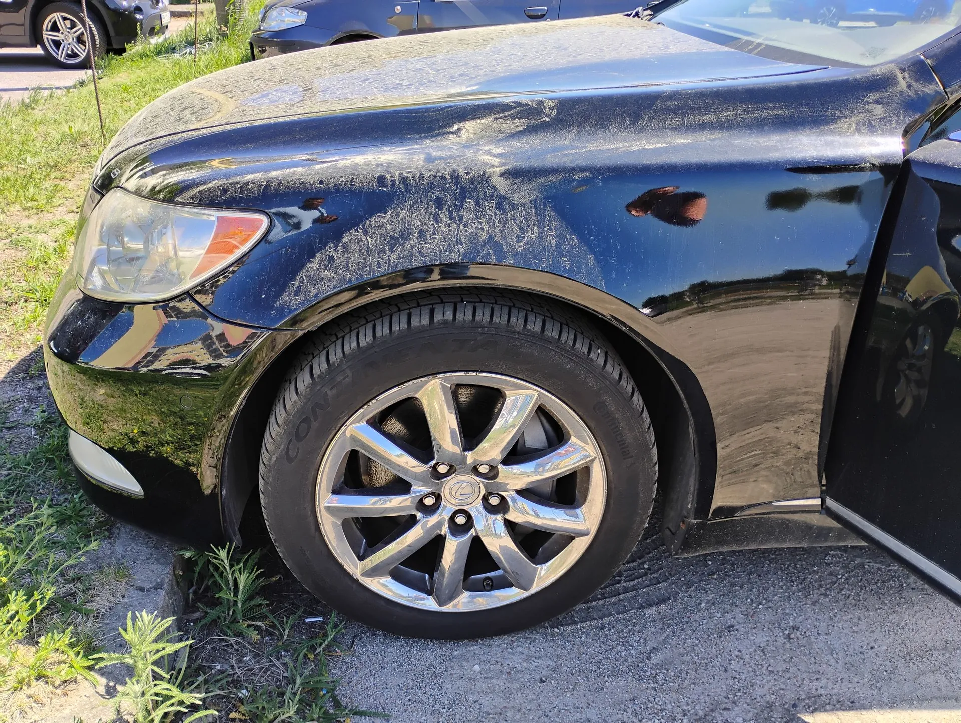 Zbliżenie na przedni lewy błotnik i koło czarnego samochodu marki Lexus. Karoseria nad kołem jest pokryta warstwą jasnego pyłu lub brudu. Widoczna jest srebrna felga z logo Lexusa w centrum oraz opona marki Continental. W tle widoczne są fragmenty innych samochodów i trawa.