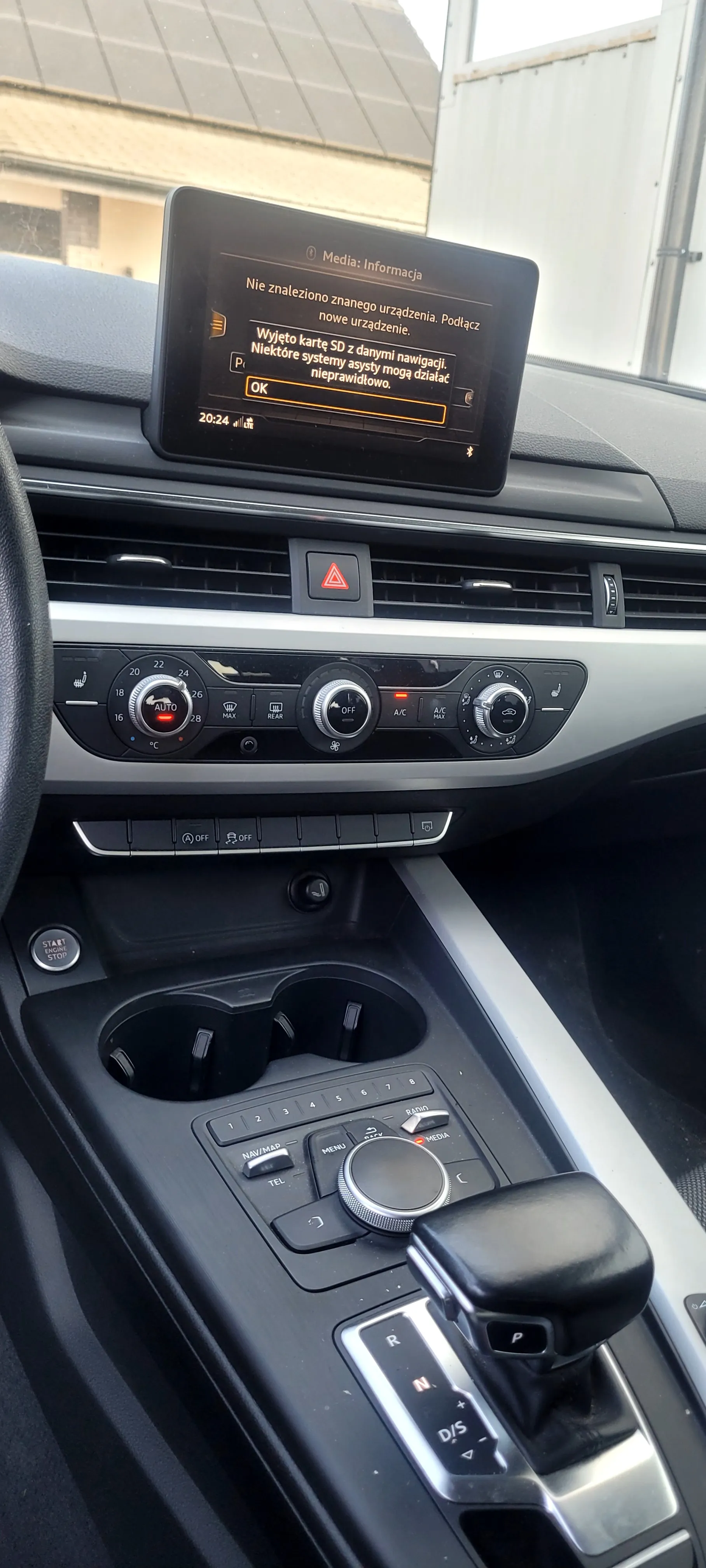 Wnętrze samochodu z widokiem na deski rozdzielczą i centralną konsolę. Na środku ekran dotykowy z napisem 'Media: Informacja. Nie znaleziono znanego urządzenia. Podłącz nowe urządzenie. Wyjęto kartę SD z danymi nawigacji. Niektóre systemy asysty mogą działać nieprawidłowo. OK' oraz godziną 20:24. Poniżej ekranu: przycisk awaryjny z czerwonym trójkątem, klimatyzacja z pokrętłami (w tym tryb AUTO), dwa uchwyty na kubki, dźwignia biegów w pozycji P (parking), przyciski NAV/MAP, TEL, MENU oraz przycisk start/stop silnika. W tle przez przednią szybę widoczny fragment budynku.