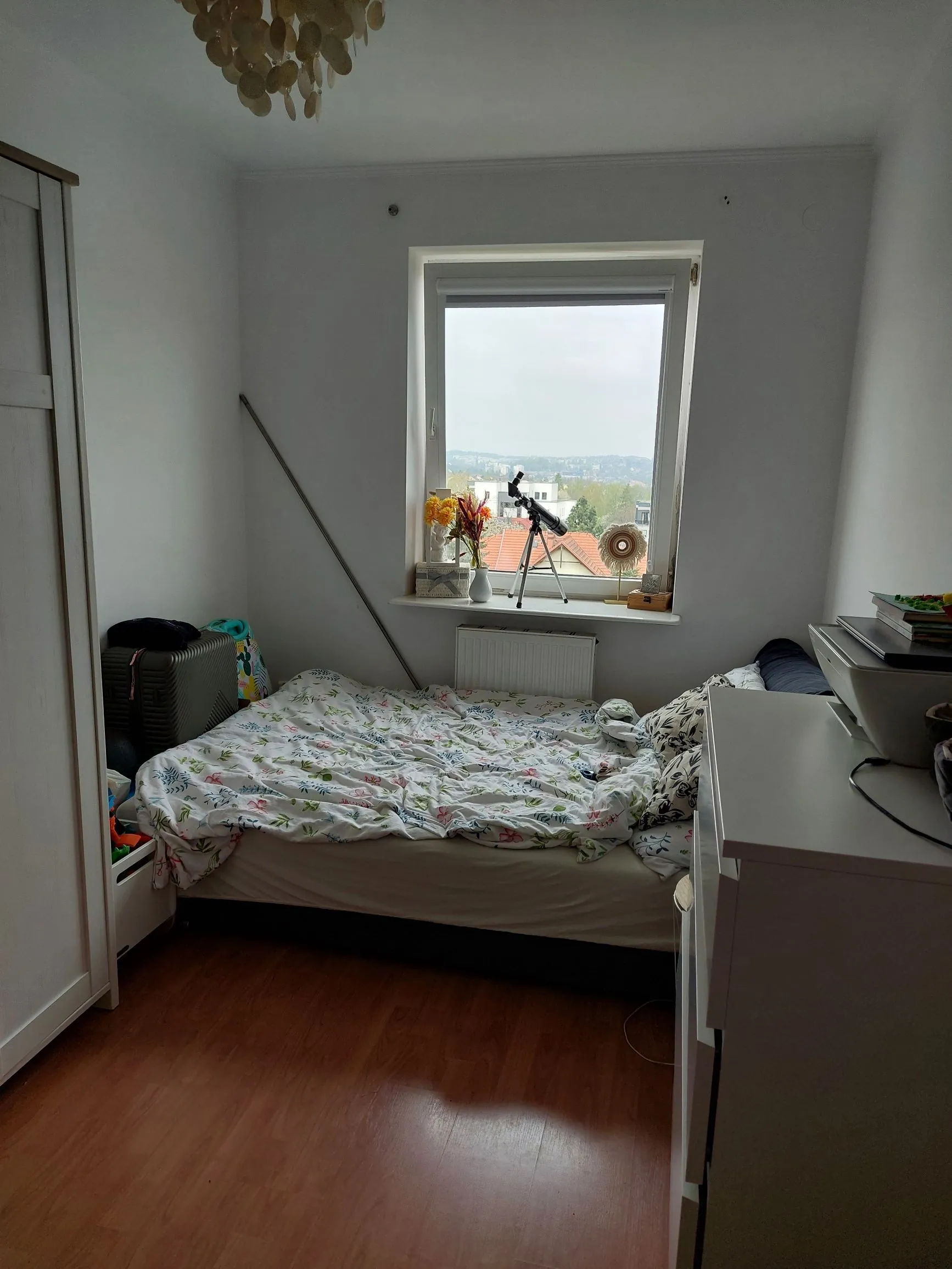 Wnętrze małego pokoju z białymi ścianami i drewnianą podłogą. W centrum znajduje się łóżko z białą pościelą w kolorowy, roślinny wzór. Nad łóżkiem jest okno, na którego parapecie stoją: wazon z żółtymi kwiatami, biały pojemnik oraz teleskop na statywie. Po lewej stronie widoczna jest biała szafa i szary plastikowy pojemnik. Po prawej stronie znajduje się biała komoda z przedmiotami na blacie. W górnej części obrazu widoczny jest fragment ozdobnej lampy.