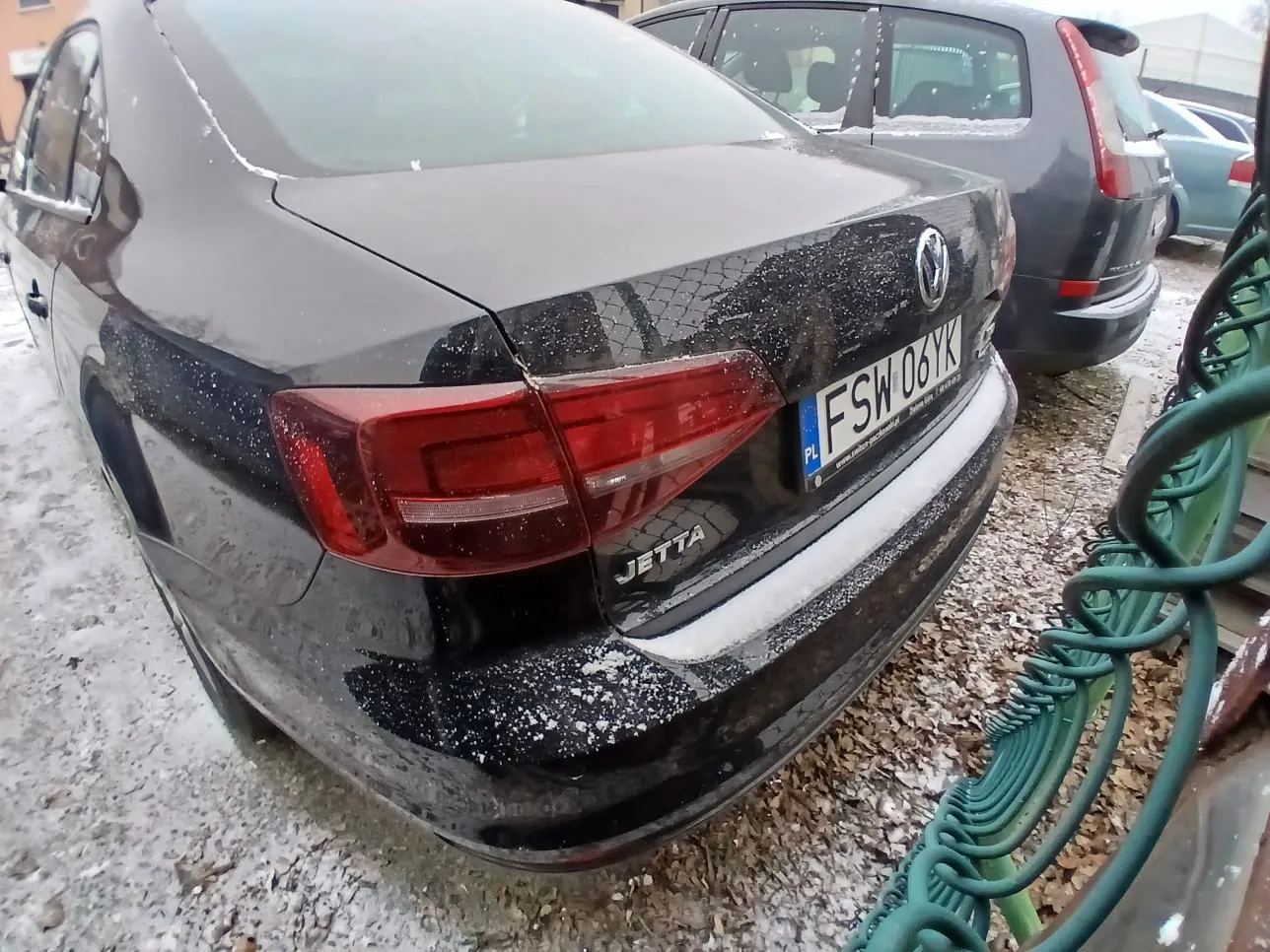Z tyłu czarnego samochodu Volkswagen Jetta widoczny jest napis JETTA oraz tablica rejestracyjna PL FSW 06YK. Na nadwoziu i szybie tylnej znajduje się śnieg. W tle zaparkowany jest szary samochód, a po prawej stronie zielone metalowe ogrodzenie. Podłoga pokryta jest śniegiem i żwirem.