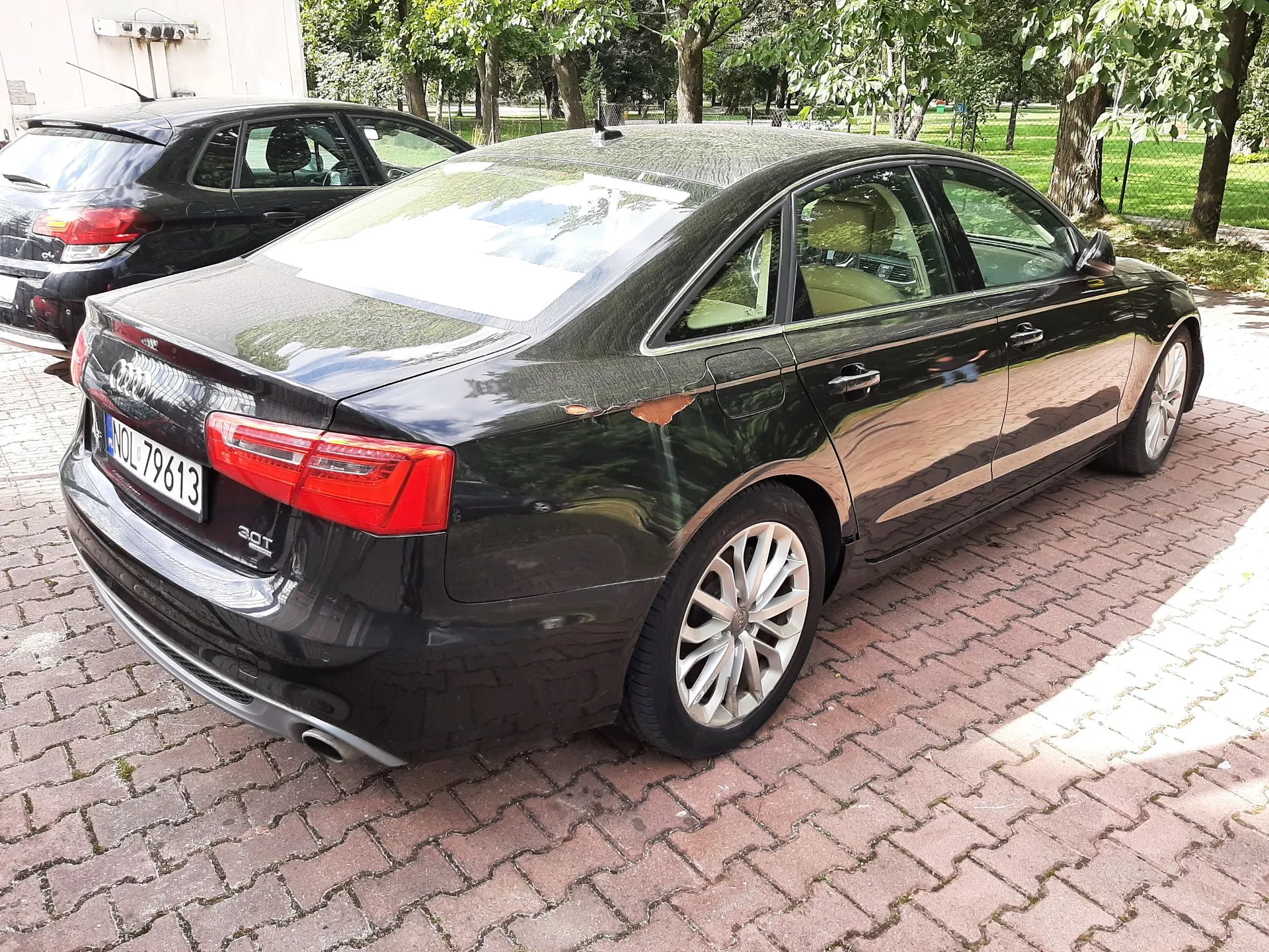 Czarny samochód marki Audi zaparkowany na kostce brukowej. Widoczny jest tył i prawy bok pojazdu. Samochód posiada polską tablicę rejestracyjną o numerze NOL 79613 oraz oznaczenie 3.0T. W tle widać fragment innego czarnego samochodu oraz zielone drzewa.