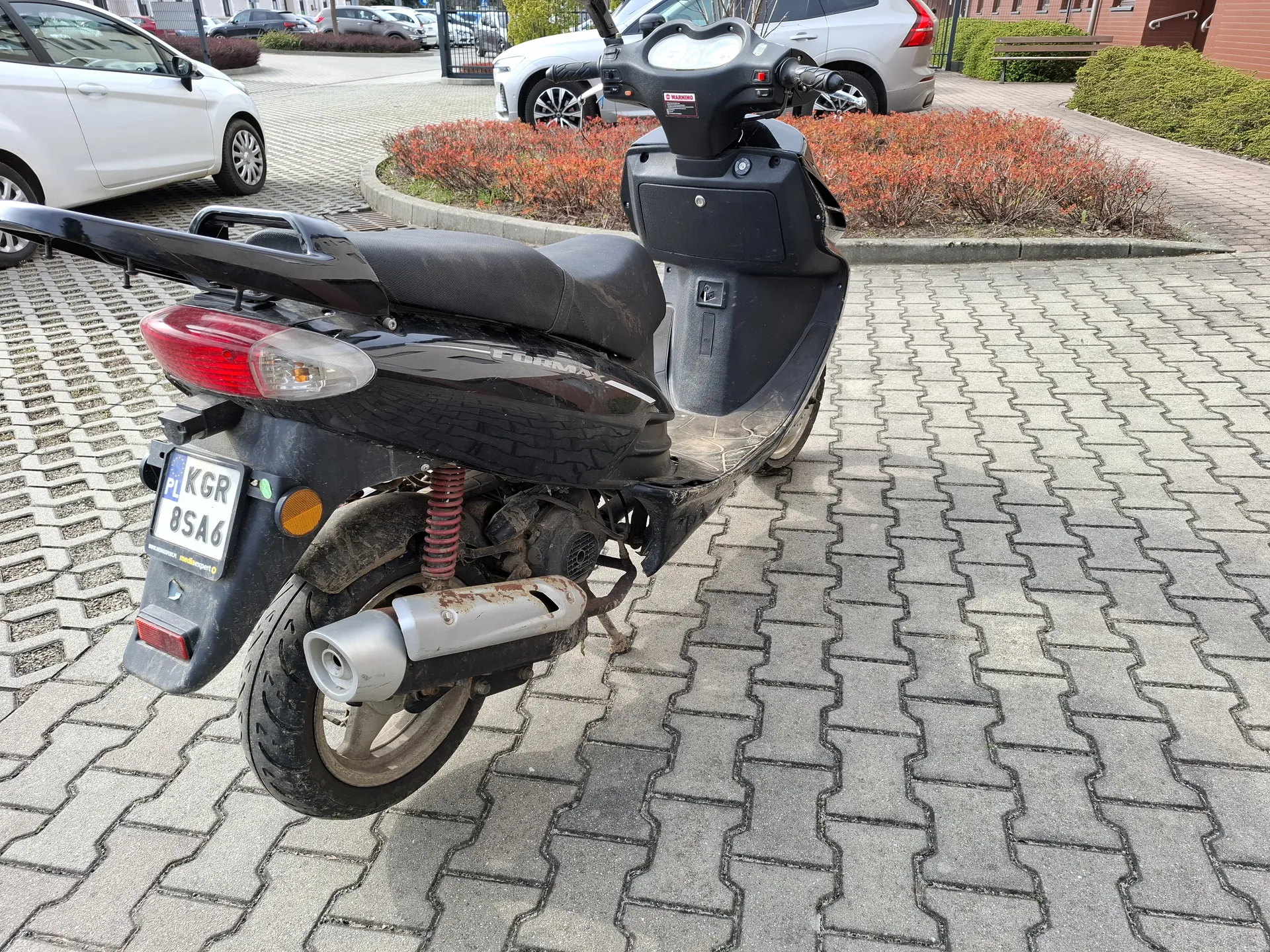 Czarny skuter zaparkowany na brukowanej powierzchni. Widoczna tablica rejestracyjna z napisem KGR 8SA6 oraz oznaczeniem PL. Skuter ma czerwone światło tylnego, pomarańczowy reflektor i zardzewiały wydech. W tle białe i srebrne samochody, krzewy z czerwonymi liśćmi oraz budynki. Tekst na tablicy rejestracyjnej jest czytelny i poziomy.