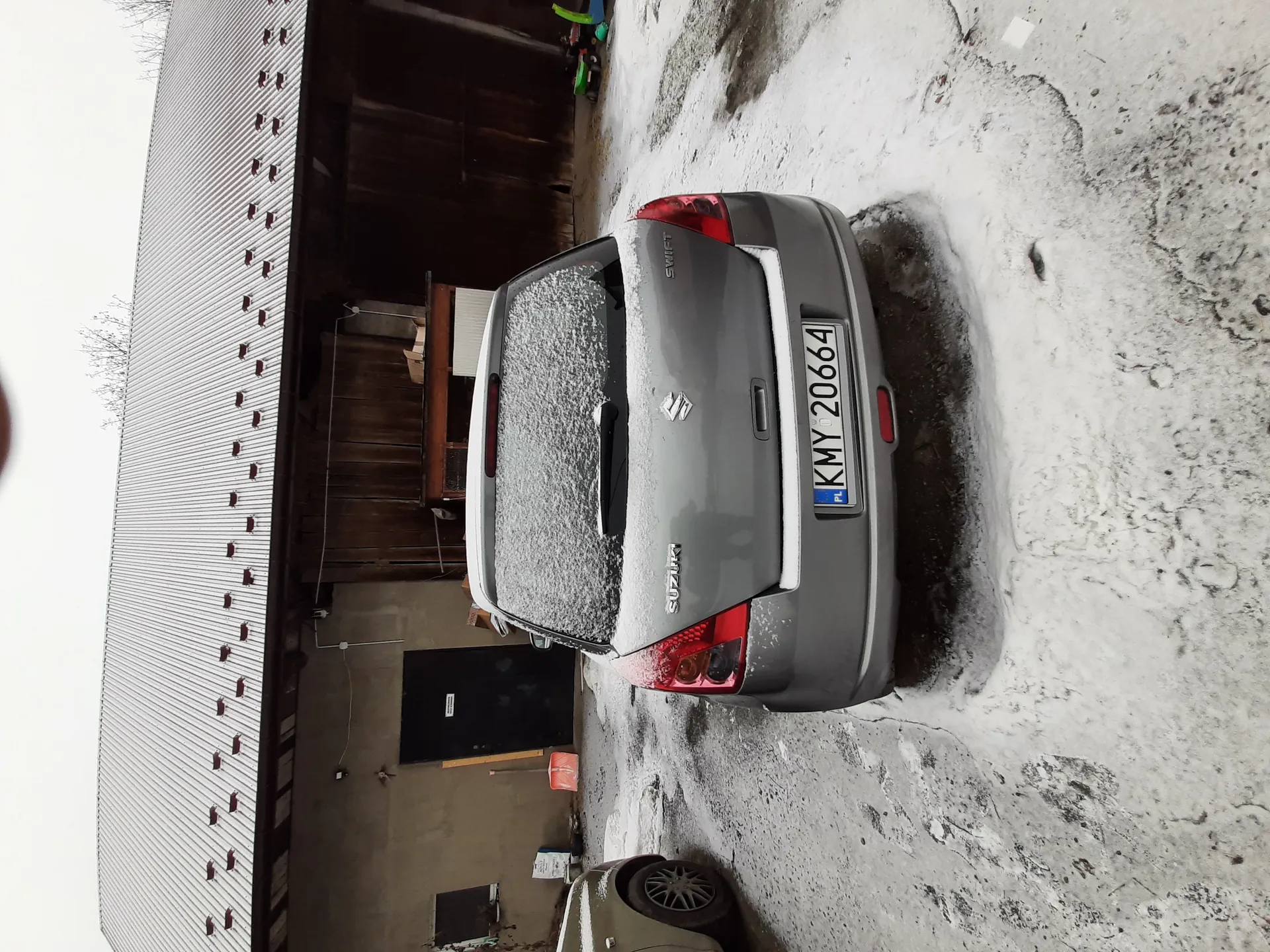 Na zdjęciu szary samochód Suzuki Swift z tablicą rejestracyjną KMY 20664 (PL), zaparkowany na nawierzchni częściowo pokrytej śniegiem. Tylne szyby i dach są śnieżne. W tle budynek z blachą falistą, otwartym wejściem z widocznymi przedmiotami i drewnianymi drzwiami. Fragment innego pojazdu.