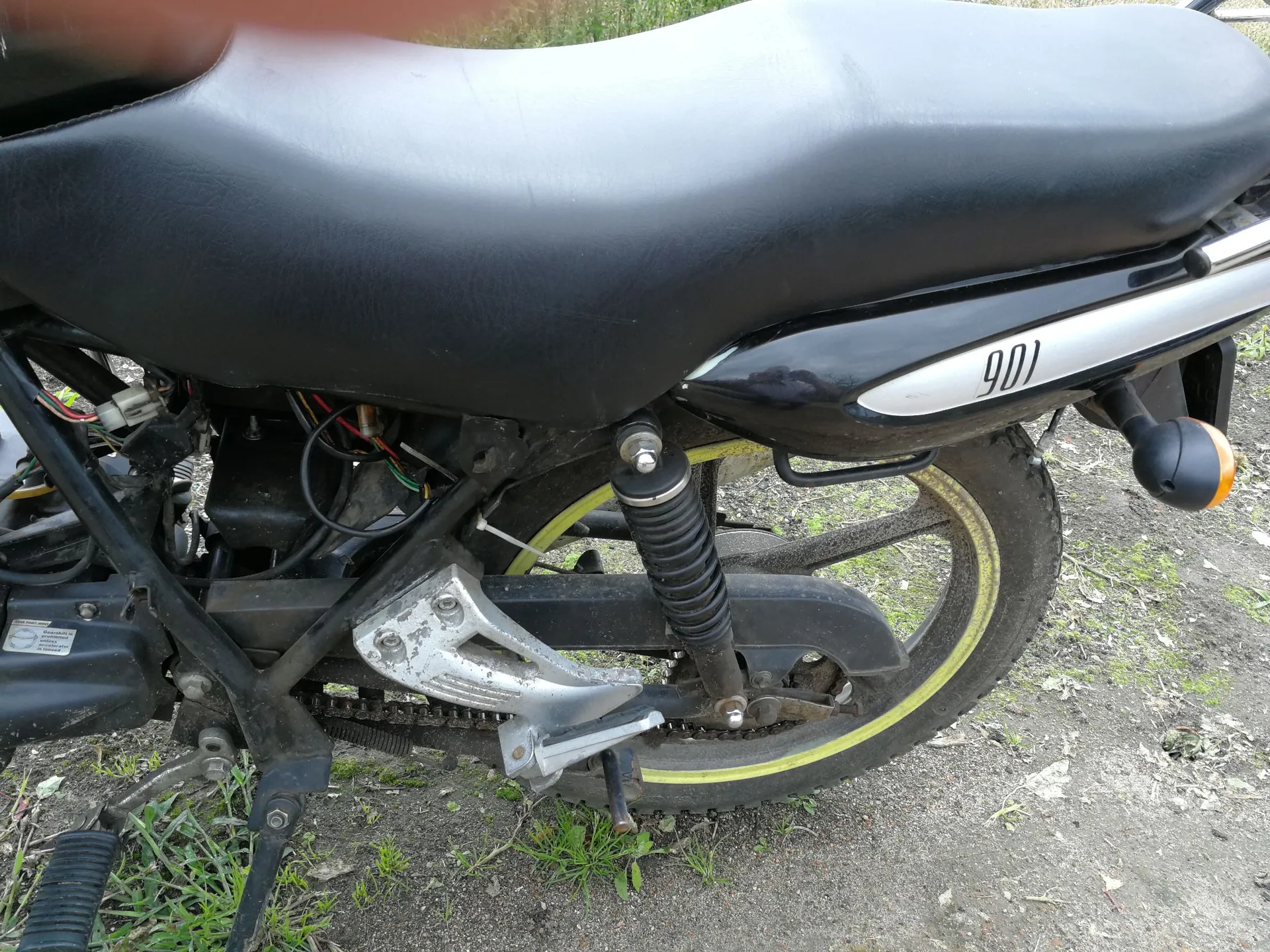 Zbliżenie na tylną część czarnego motocykla. Widoczne jest czarne, skórzane siedzenie, tylne koło z felgą z żółtym paskiem, amortyzator oraz łańcuch napędowy. Na bocznej owiewce znajduje się biały napis '901'. Motocykl stoi na piaszczystym podłożu z kępkami trawy.