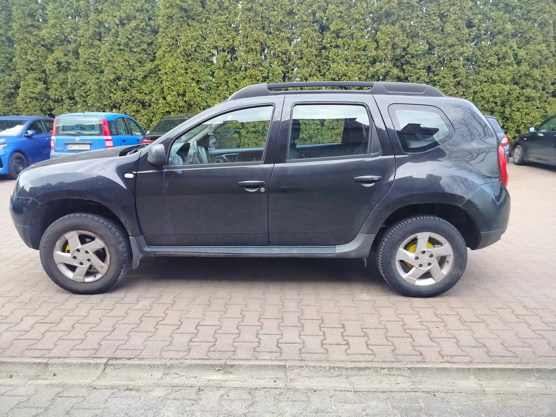Ciemnoszary SUV z widocznym profilem bocznym, dachowymi przenośnikami i srebrnymi felgami z żółtymi akcentami. W tle niebieski samochód z tablicą rejestracyjną WPZ 44370 oraz inne pojazdy. Tło stanowią zielone drzewa, a podłoga to brukowana powierzchnia parkingowa z chodnikiem.
