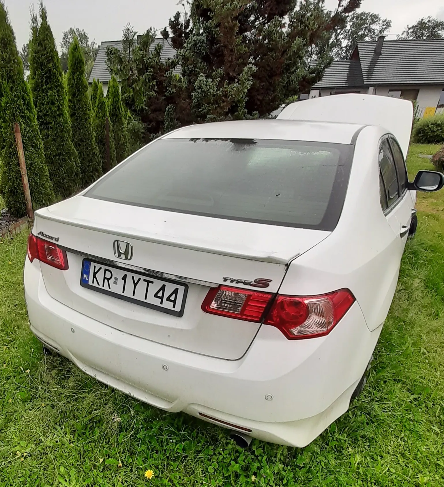 Tylny widok białego samochodu marki Honda Accent z otwartą maską przednią, zaparkowanego na zielonej trawie. Samochód posiada polską tablicę rejestracyjną o numerze KR 1YT44 oraz oznaczenie Type S na klapie bagażnika. W tle widoczne są wysokie, zielone drzewa iglaste oraz dachy domów jednorodzinnych.