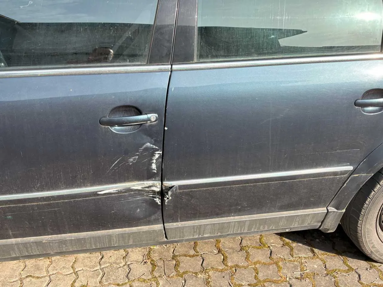 Boczny widok ciemnoszarego samochodu z widocznymi uszkodzeniami na drzwiach: wgnieceniem i zadrapaniami. Samochód stoi na brukowanej powierzchni z kamieni w kształcie sześciokątów. Widoczne są czarne klamki drzwi, przyciemniane szyby oraz fragment przedniego koła. W odbiciu na szybie widoczna sylwetka osoby trzymającej aparat.