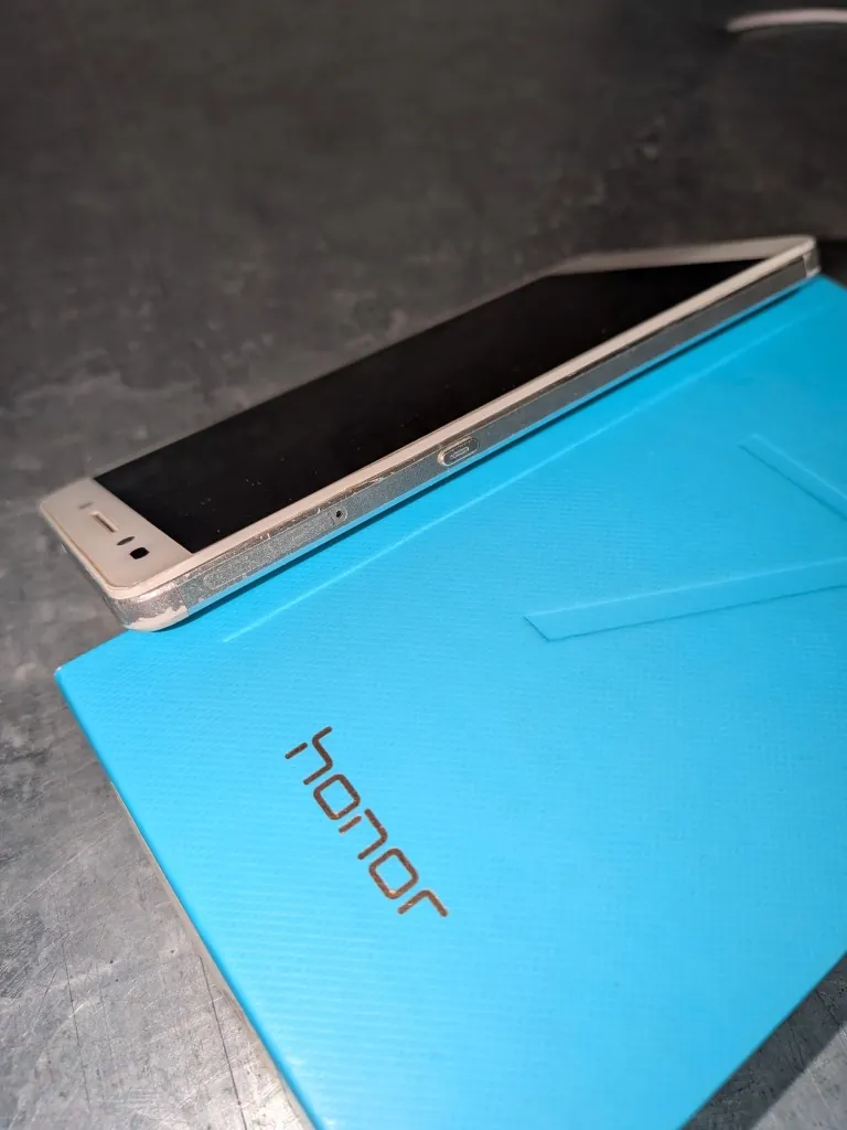 Smartfon z białymi ramkami leżący na niebieskim etui z napisem 'honor'. Urządzenie jest widoczne z perspektywy bocznej, ukazując krawędź obudowy z widocznymi śladami zużycia i zarysowaniami. Całość spoczywa na ciemnej, teksturowanej powierzchni.