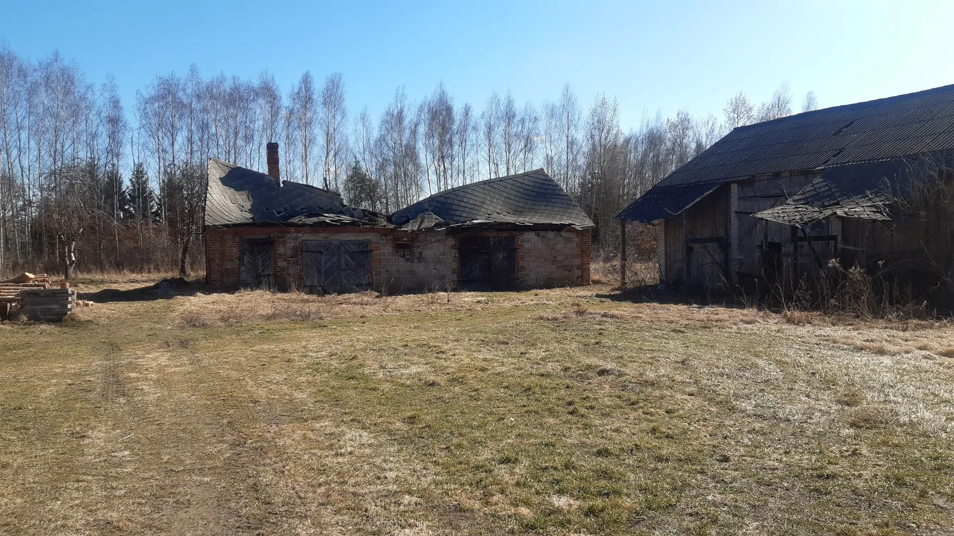 Zdjęcie przedstawiające dwa zniszczone, stare budynki gospodarcze na tle lasu z bezlistnymi drzewami. Budynek po lewej jest częściowo zburzony, ma cegiane ściany i zapadnięty dach pokryty ciemną papą. Budynek po prawej jest większy, wykonany z ciemnego drewna z dwuspadowym dachem. Przed budynkami znajduje się obszar z wyschniętą, żółtawą trawą i ziemistą drogą. Niebo jest bezchmurne i jasnoniebieskie.
