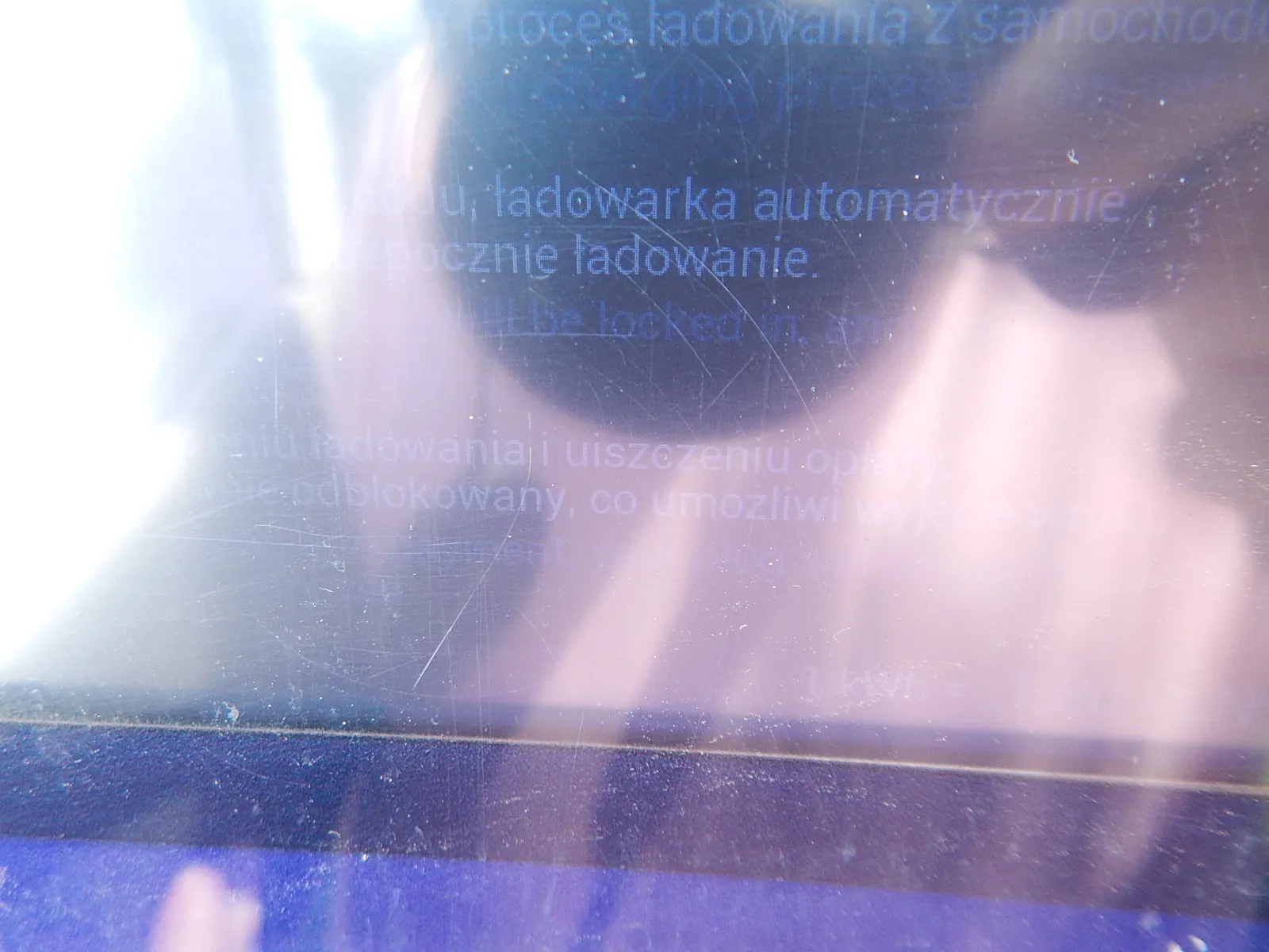 Zbliżenie na powierzchnię z tekstem w języku polskim, która jest mocno prześwietlona i zarysowana. Widoczne fragmenty zdań: 'ładowania z samochodu', 'ładowarka automatycznie rozpocznie ładowanie' oraz 'odblokowany, co umożliwi wyjazd'. Tekst jest częściowo nieczytelny z powodu odblasków światła.