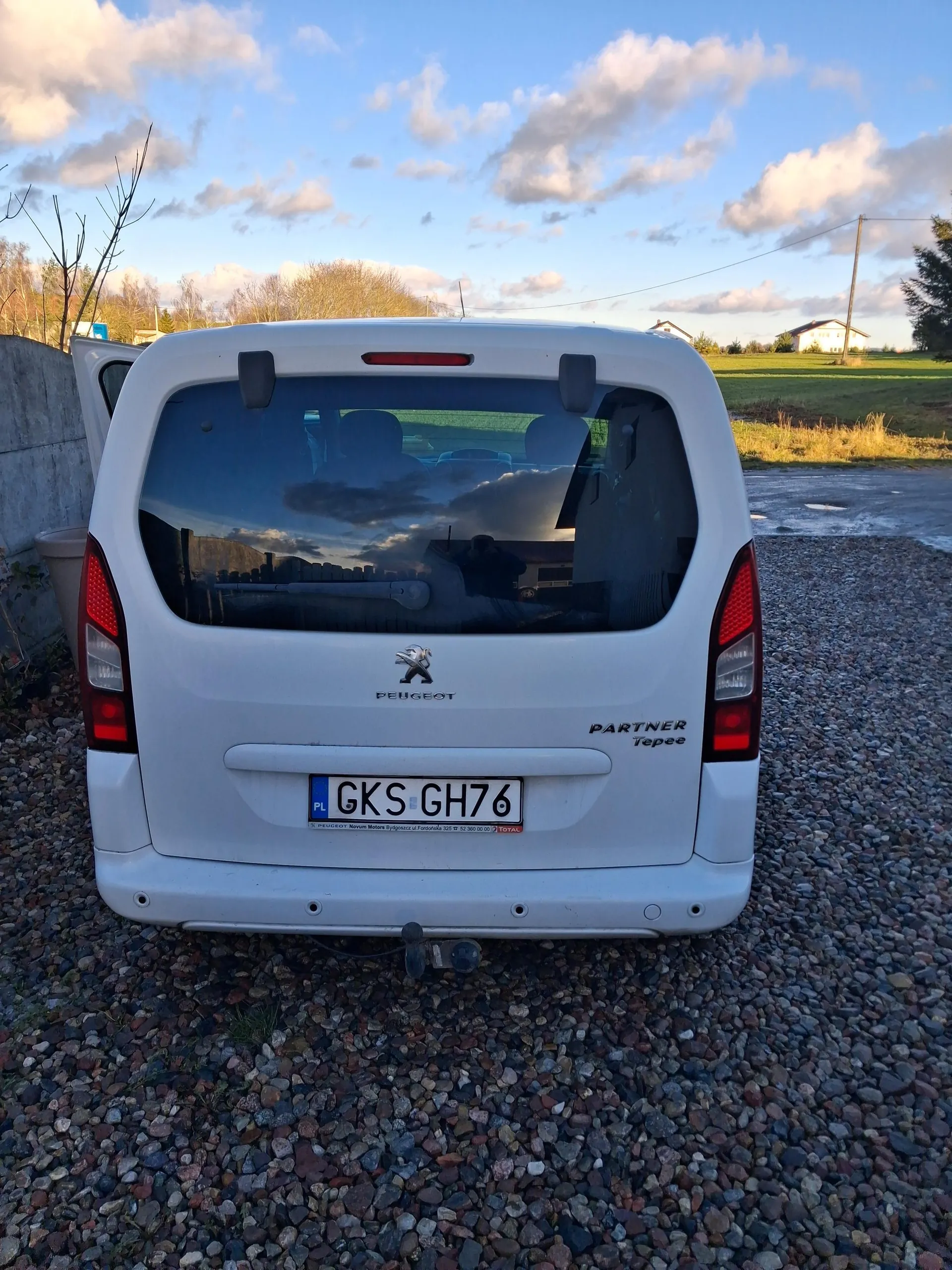 Biały samochód dostawczy Peugeot Partner Tepee z widocznym tyłem. Na tablicy rejestracyjnej widnieje numer GKS GH76 z kodem kraju PL. Samochód stoi na żwirowym podłożu, z lewej strony otwarte drzwi. W tle betonowa ściana, drzewa, domy i niebo z chmurami. Na tylnym szybie odbicie chmur i otoczenia.