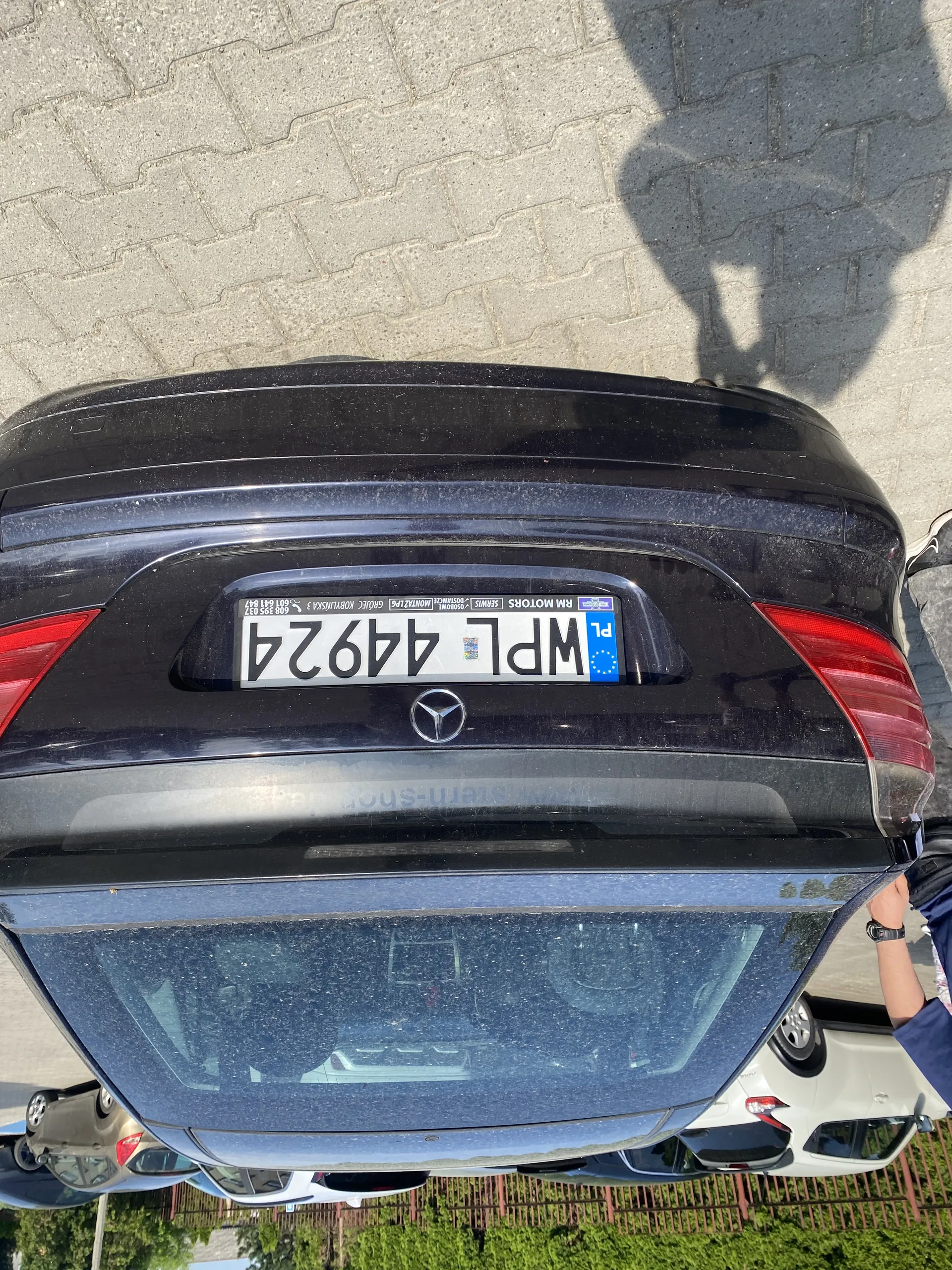 Tył czarnego samochodu marki Mercedes-Benz z tablicą rejestracyjną WPL 44924. Na tablicy widoczne są oznaczenia 'PL' z flagą Unii Europejskiej oraz napis 'RM MOTORS'. W tle inne zaparkowane samochody, brukowana powierzchnia, zieleń i ogrodzenie. Widoczna również ręka osoby w niebieskiej koszulce.