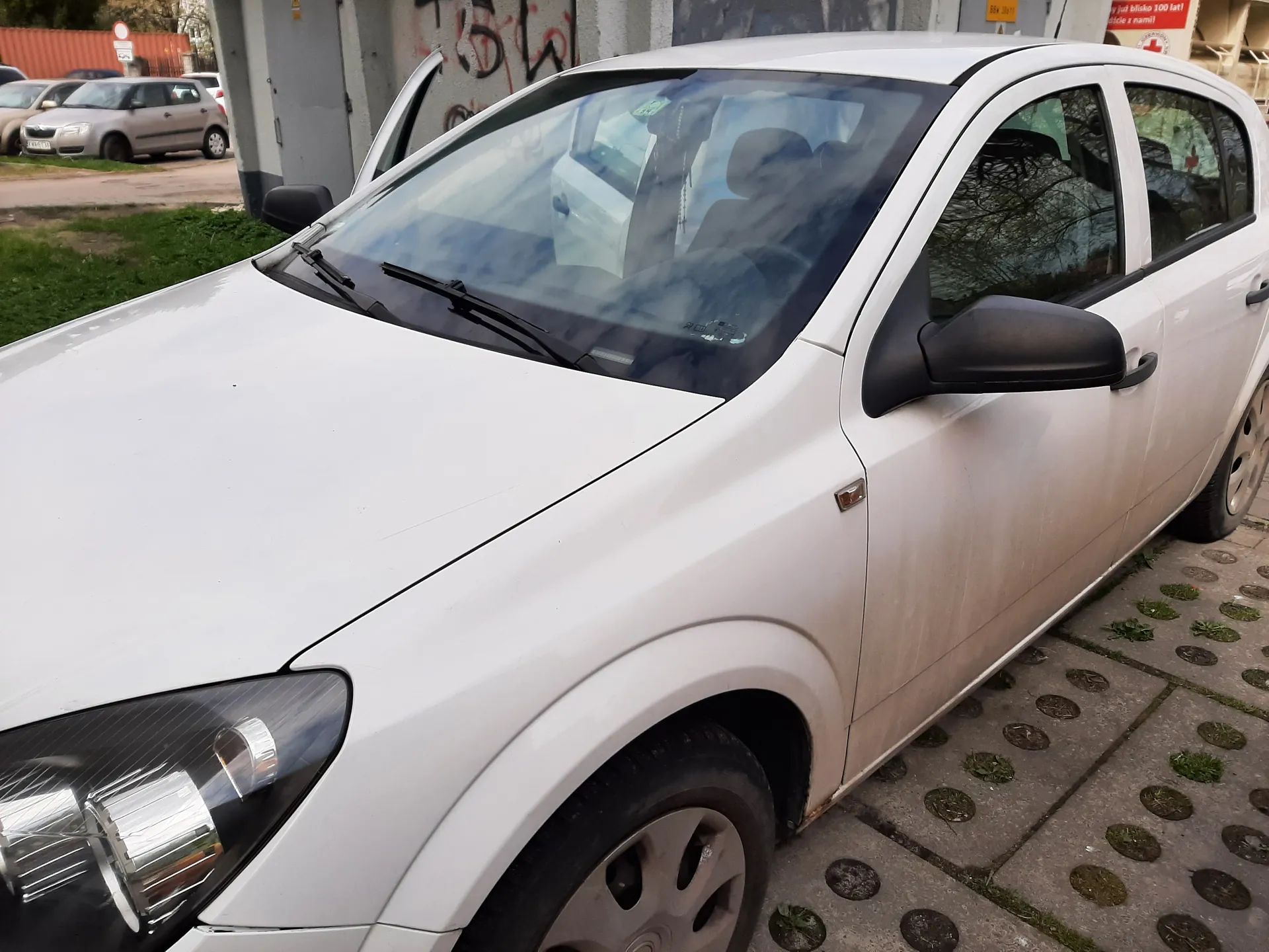Biały samochód osobowy zaparkowany na chodniku z okrągłymi betonowymi płytami i trawą między nimi. Drzwi przednie po lewej stronie są otwarte. Na tle widoczne są inne zaparkowane samochody, betonowa ściana z graffiti oraz czerwony znak z białym tekstem: 'Ból jest blisko 100 lat! ... z nami!'. Samochód ma lekko zabrudzoną karoserię, zwłaszcza dolne części drzwi. W tle również zielona trawa i fragment budynku.