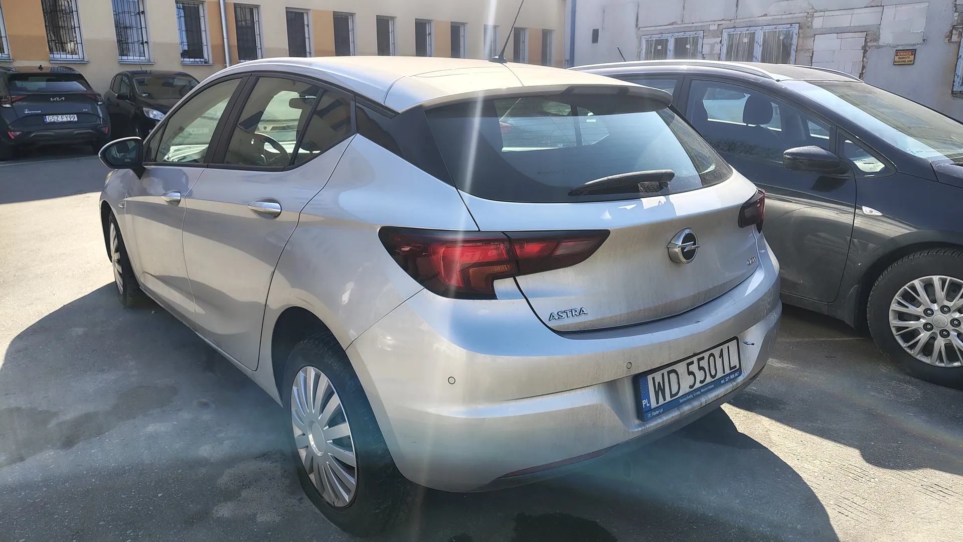 Srebrny samochód Opel Astra widziany od tyłu, z tablicą rejestracyjną WD 5501L. Na tle budynki z oknami, inne zaparkowane pojazdy (w tym czarny samochód Kia z tablicą G57 FY99) oraz asfaltowy parking. Na tylnym zderzaku widoczny napis Astra i logo marki. Słoneczny dzień, cienie na ziemi.