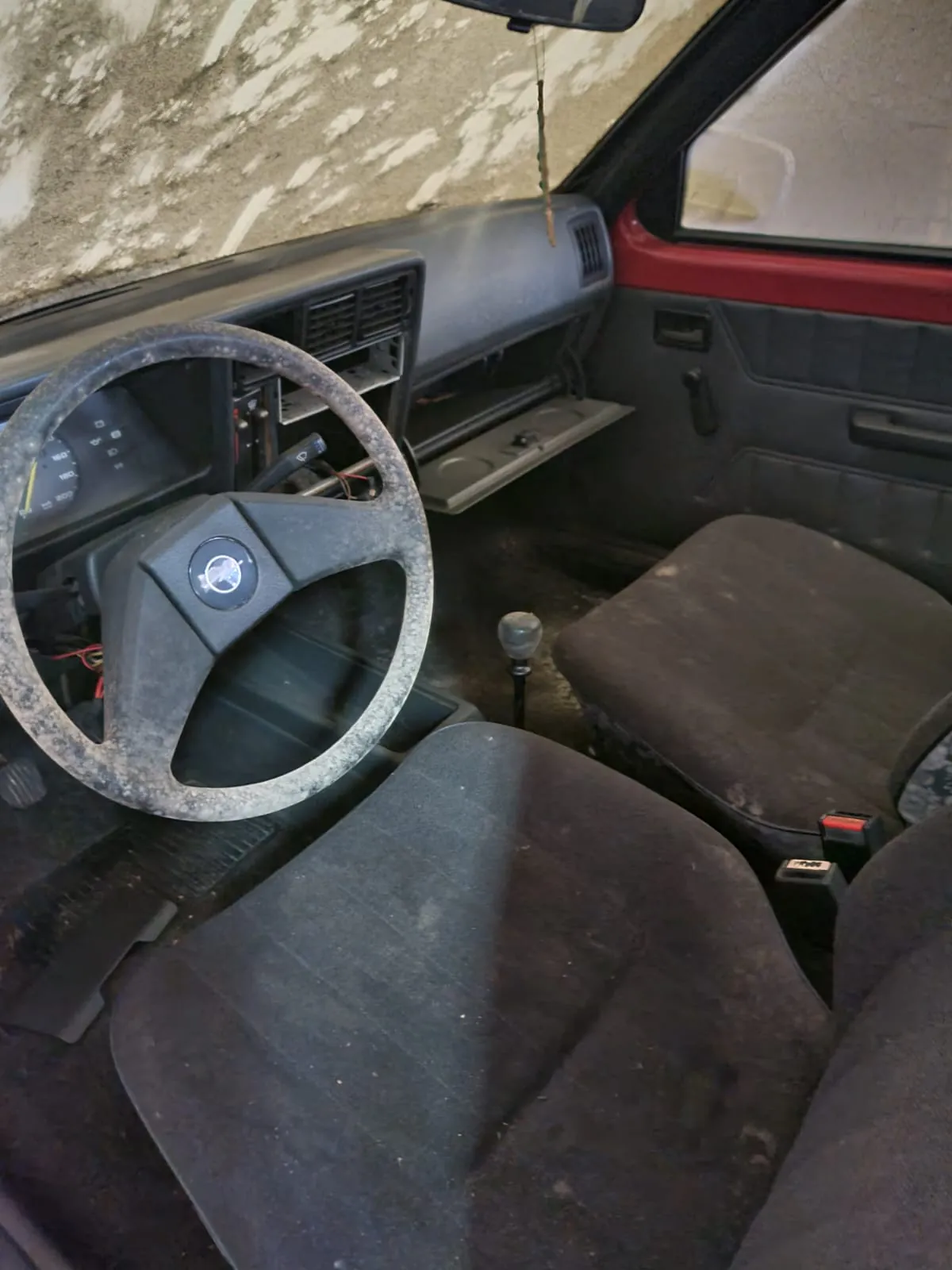 Wnętrze starego samochodu z kierownicą po lewej stronie, zniszczonymi siedzeniami pokrytymi kurzem, deską rozdzielczą z otwartymi przestrzeniami, dźwignią zmiany biegów, czerwoną obudową drzwi i betonową ścianą z uszkodzoną sztukатурą widoczną przez przednią szybę.