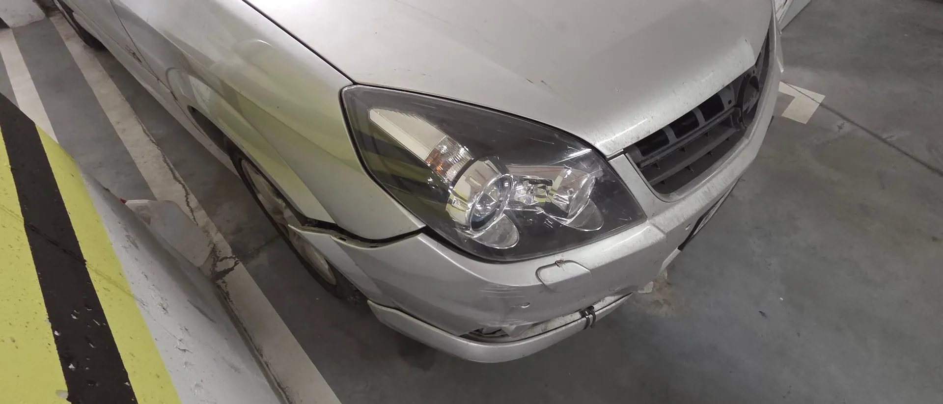 Zbliżenie na przedni prawy róg srebrnego samochodu. Widoczny jest reflektor, grill oraz uszkodzony zderzak z wgnieceniami i zarysowaniami w miejscu łączenia z błotnikiem. Samochód stoi na betonowym parkingu obok żółto-czarnego oznaczenia na ziemi.