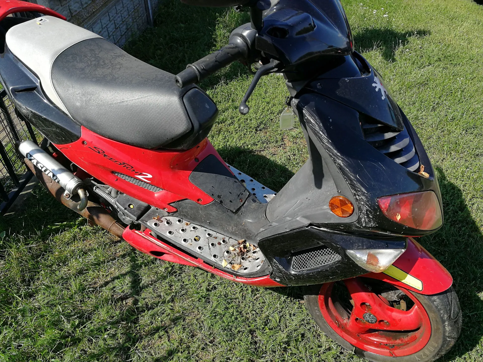 Czarny i czerwony skuter zaparkowany na trawie. Pojazd ma czarną przednią owiewkę, czerwone boki z napisem 'Sportline 2' oraz czerwone felgi. Widoczne jest srebrne wydech z napisem 'VASUNI'. Skuter jest zakurzony i widać na nim ślady użytkowania.