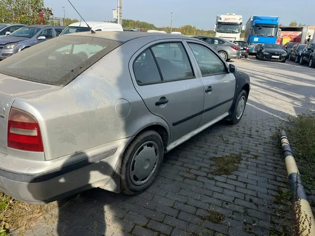 Srebrny samochód osobowy typu hatchback zaparkowany na kostce brukowej. Pojazd jest zakurzony, widoczne są zarysowania na boku oraz uszkodzony zderzak tylny. W tle znajduje się parking z innymi samochodami oraz ciężarówkami, w tym niebieska ciężarówka po prawej stronie.