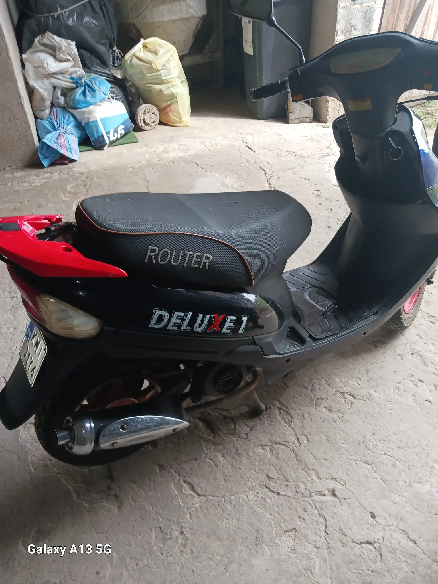 Czarny skuter z czerwonymi akcentami na tle betonowej podłogi. Na siedzeniu widnieje napis 'ROUTER', a na boku skutera 'DELUXE 1' z czerwonym 'X'. W tle stos worków (żółty, niebiesko-biały, czarny) oraz betonowa konstrukcja. Tablica rejestracyjna z częściowo widocznym numerem. W lewym dolnym rogu napis 'Galaxy A13 5G'.