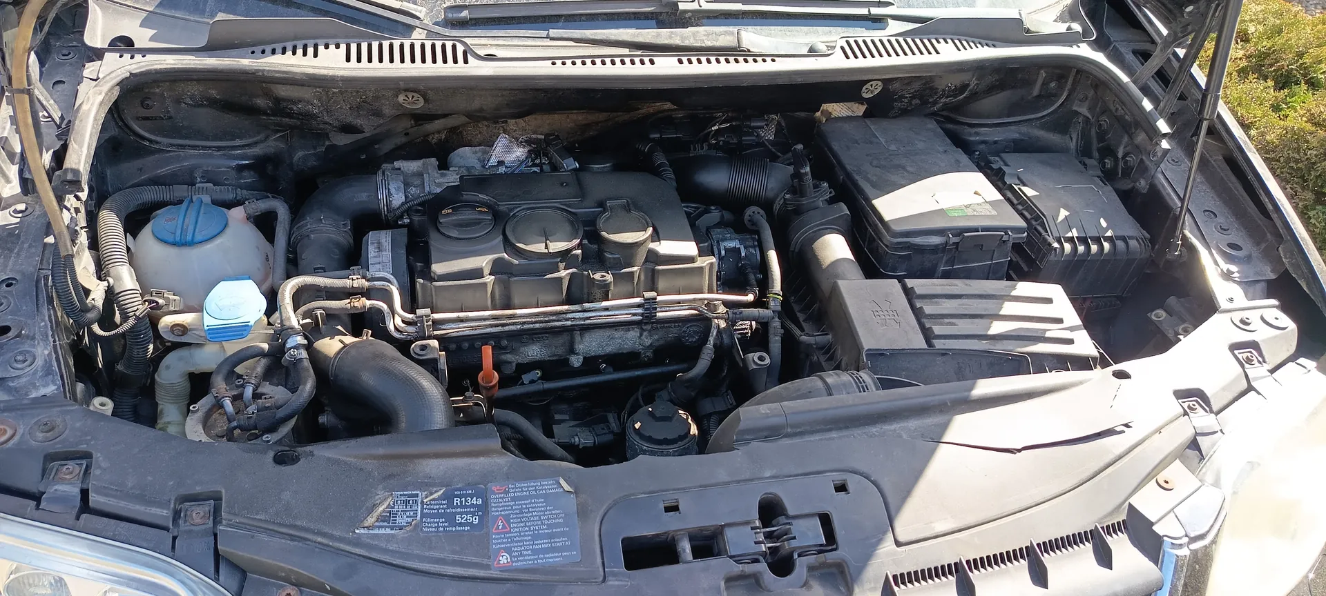 Zdjęcie przedstawia otwarty komorę silnika samochodu. Widać elementy takie jak zbiornik chłodniczy z niebieską pokrywką, różne przewody, czarne osłony oraz etykietę z napisami 'R134a' i '525g'. Samochód stoi na zewnątrz, w tle widoczna jest trawa. Komora silnika jest czysta, a elementy są ułożone w typowej konfiguracji.