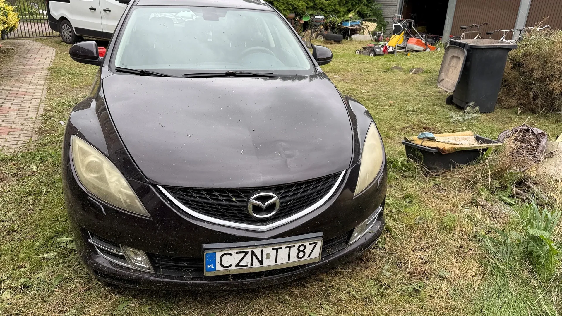 Widok z przodu na ciemnofioletowy samochód marki Mazda zaparkowany na trawie. Pojazd ma polską tablicę rejestracyjną o numerze CZN TT87. W tle widoczny jest biały samochód dostawczy, trawnik oraz różne przedmioty gospodarcze, w tym czarny kosz na śmieci i kosiarka.