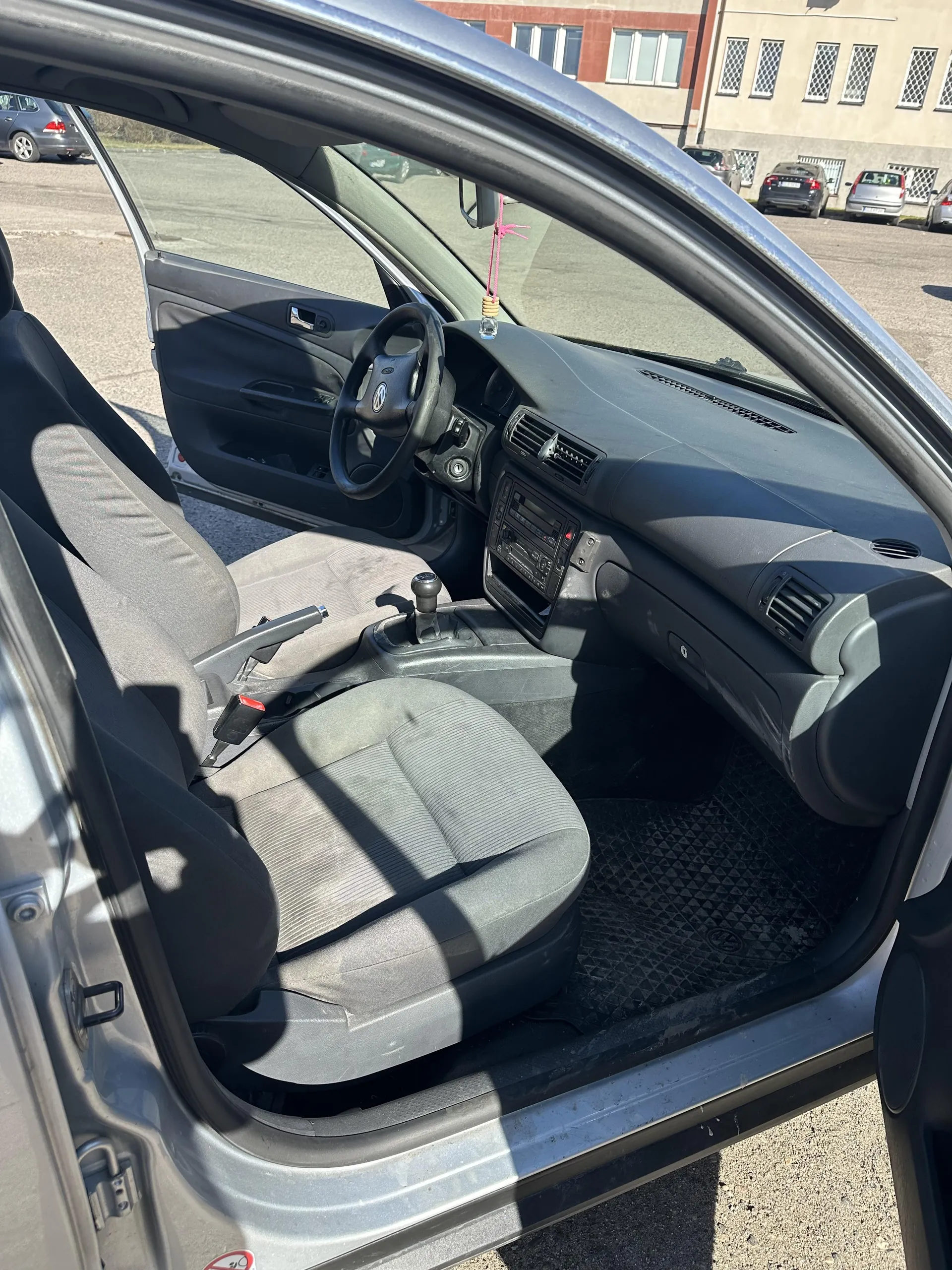 Wnętrze samochodu z otwartymi drzwiami od strony kierowcy. Widoczne są szare tkaninowe fotele, kierownica z logo Volkswagen, deska rozdzielcza z radioodtwarzaczem i wentylacjami, dźwignia zmiany biegów oraz dywanik z logo VW. Na zewnątrz widać parking z innymi pojazdami i budynkiem o czerwono-beżowej elewacji z oknami. Na lusterku wstecznym wiszą różowe ozdoby.