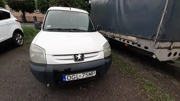 Biały samochód dostawczy marki Peugeot z tablicą rejestracyjną OGL 75MP, stojący na podwórku z żwirem i trawą. Po lewej stronie częściowo widoczny biały samochód osobowy, po prawej stronie przyczepa z ciemnym pokryciem. W tle drzewa i fragment budynku.