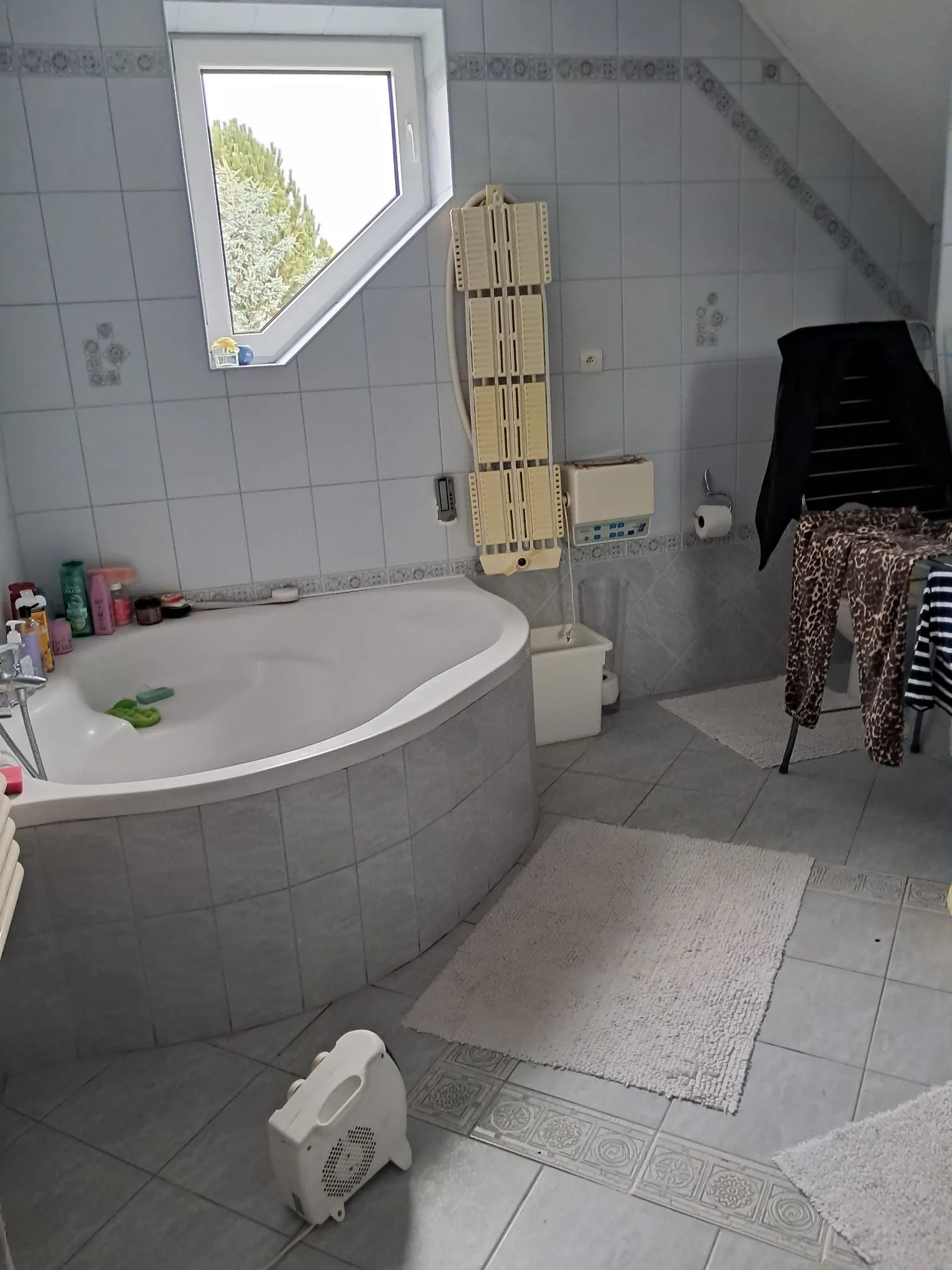 Łazienka z białą wanną w narożniku, obłożoną szarymi płytkami. Na brzegu wanny znajdują się kolorowe butelki z kosmetykami. W tle widać białe kafelki na ścianach, okno dachowe z widokiem na zieleń oraz beżowy grzejnik drabinkowy. Obok grzejnika stoi biały kosz na śmieci. Po prawej stronie znajduje się czarny wieszak z ubraniami, w tym spodniami w panterkę. Na podłodze leżą jasne dywaniki łazienkowe, a w dolnej części obrazu widoczny jest biały grzejnik elektryczny.