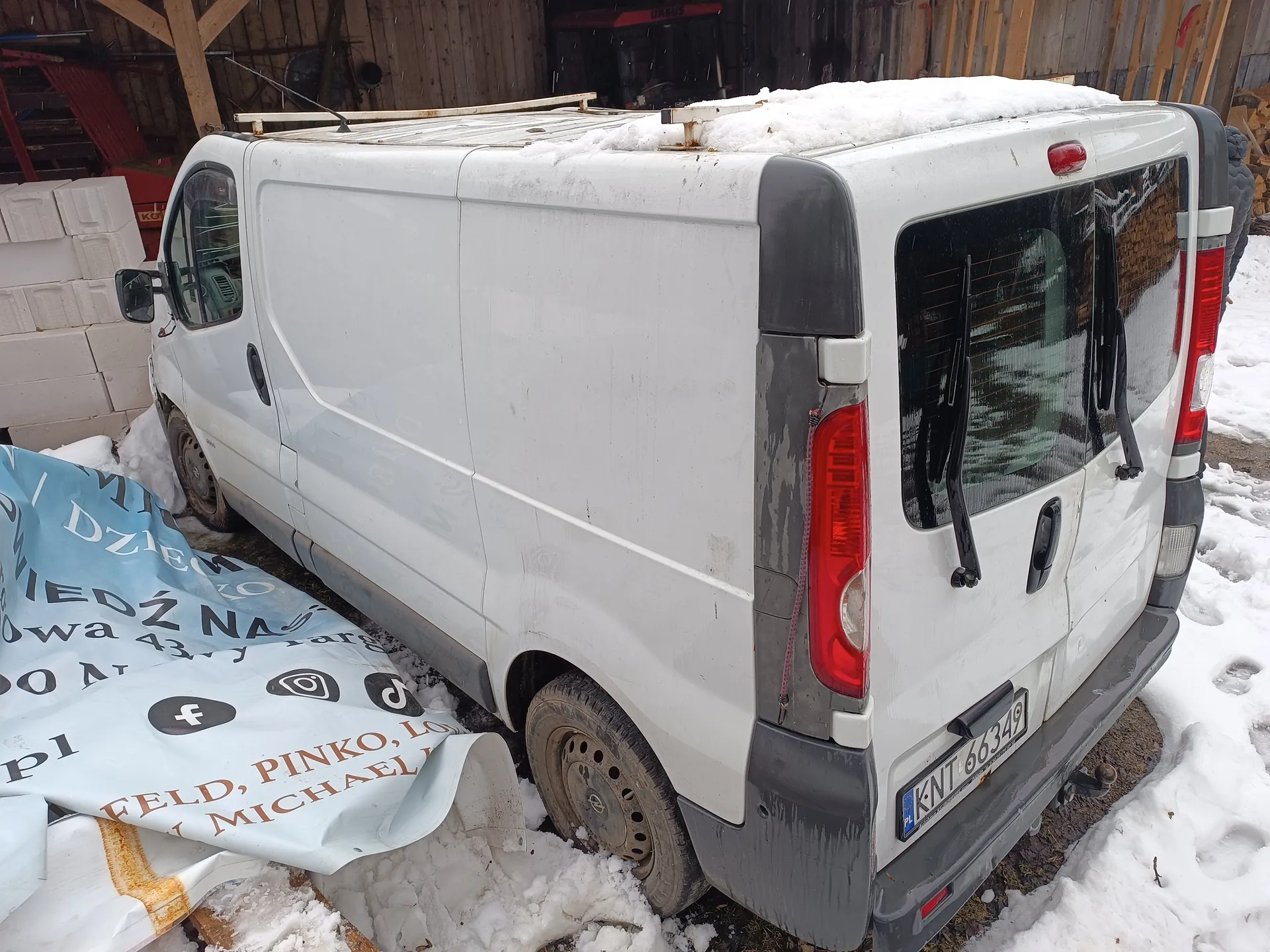 Biały furgon z tablicą rejestracyjną KNT 66349, pokryty śniegiem na dachu, zaparkowany na śnieżnej powierzchni. Obok furgona leży niebieska folia z widocznym tekstem: DZIECI, NIEDŹWIA, 43, FELD, PINKO, LO, MICHAEL oraz ikonami mediów społecznościowych (Facebook, TikTok). W tle drewniana budowla, białe bloki i czerwony traktor. Furgon ma szare elementy na zderzaku i krawędziach, czerwone światła tylnego oraz wycieraczki na tylnych szybach.