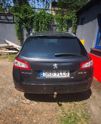 Tylny widok ciemnoszarego samochodu marki Peugeot 508 SW zaparkowanego na piaszczystym podłożu. Samochód ma polską tablicę rejestracyjną o numerze SRB 9LL6. W tle widoczny jest szary mur porośnięty zielonymi roślinami oraz fragment niebieskiego budynku po prawej stronie.