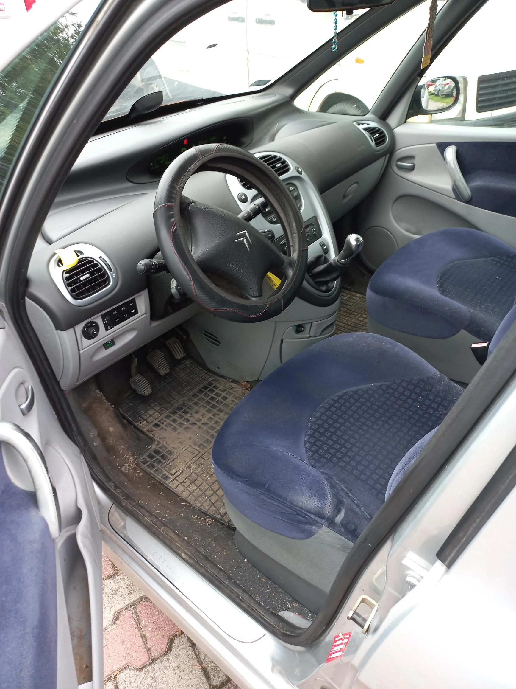 Wnętrze samochodu widziane z otwartych drzwi kierowcy. Widoczna jest szara deska rozdzielcza, czarna kierownica z logo Citroëna, dwa przednie fotele obite ciemnoniebieskim materiałem oraz pedały w przestrzeni dla nóg. Na desce rozdzielczej znajdują się otwory nawiewów i panel sterowania.
