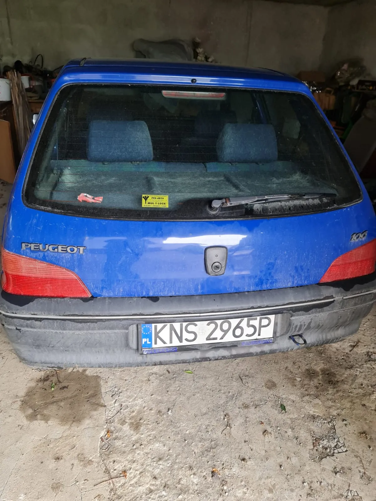 Z tyłu niebieski samochód marki PEUGEOT 106 z tablicą rejestracyjną KNS 2965P (PL). Na tylnej szybie widoczny żółty naklejka oraz czerwony element dekoracyjny. Samochód stoi w pomieszczeniu z betonowymi ścianami i podłogą, na której znajdują się plamy i śmieci. W tle widoczne narzędzia i przedmioty gospodarstwa domowego.