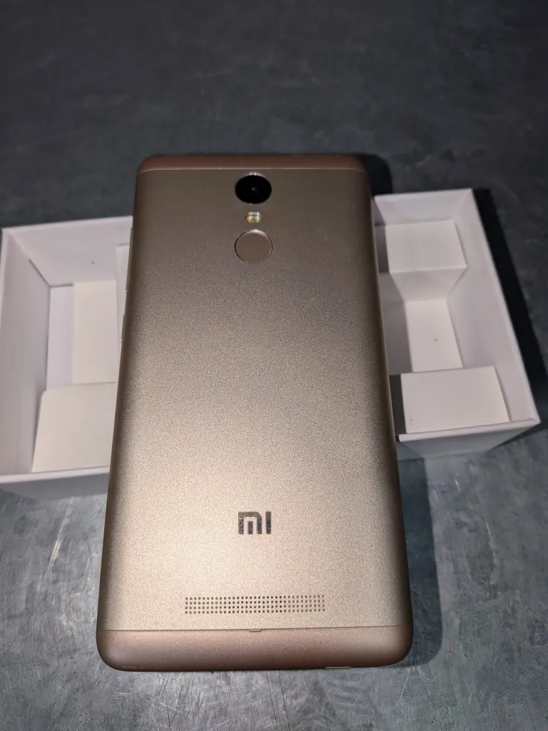 Tylna obudowa złotego smartfona marki Xiaomi z logo 'mi' na dole. Na górze urządzenia znajduje się aparat fotograficzny, lampa błyskowa oraz czytnik linii papilarnych. Telefon leży na szarym, metalicznym blacie, częściowo w białym kartonowym opakowaniu.