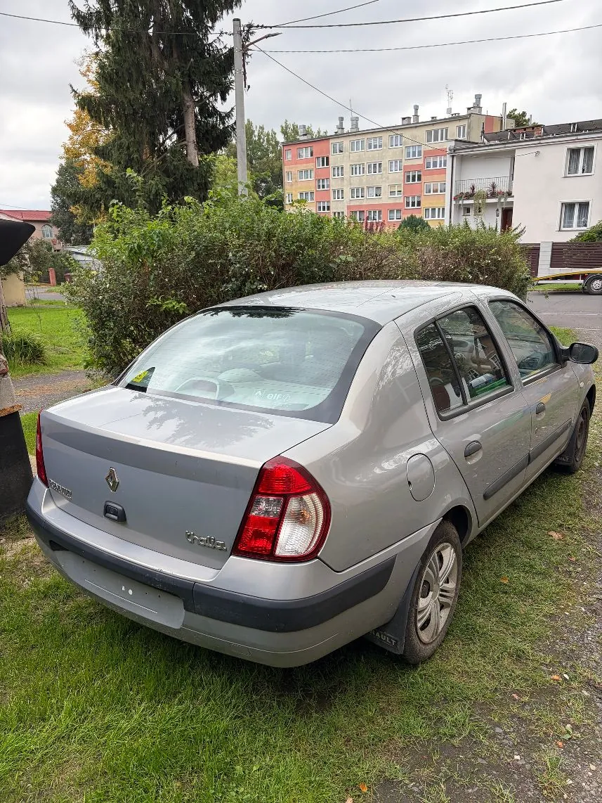 Srebrny samochód marki Renault Mégane zaparkowany na trawiastym poboczu. Widok z tyłu i z boku pojazdu. W tle znajdują się zielone krzewy oraz wielopiętrowy blok mieszkalny w kolorach białym i pomarańczowym.