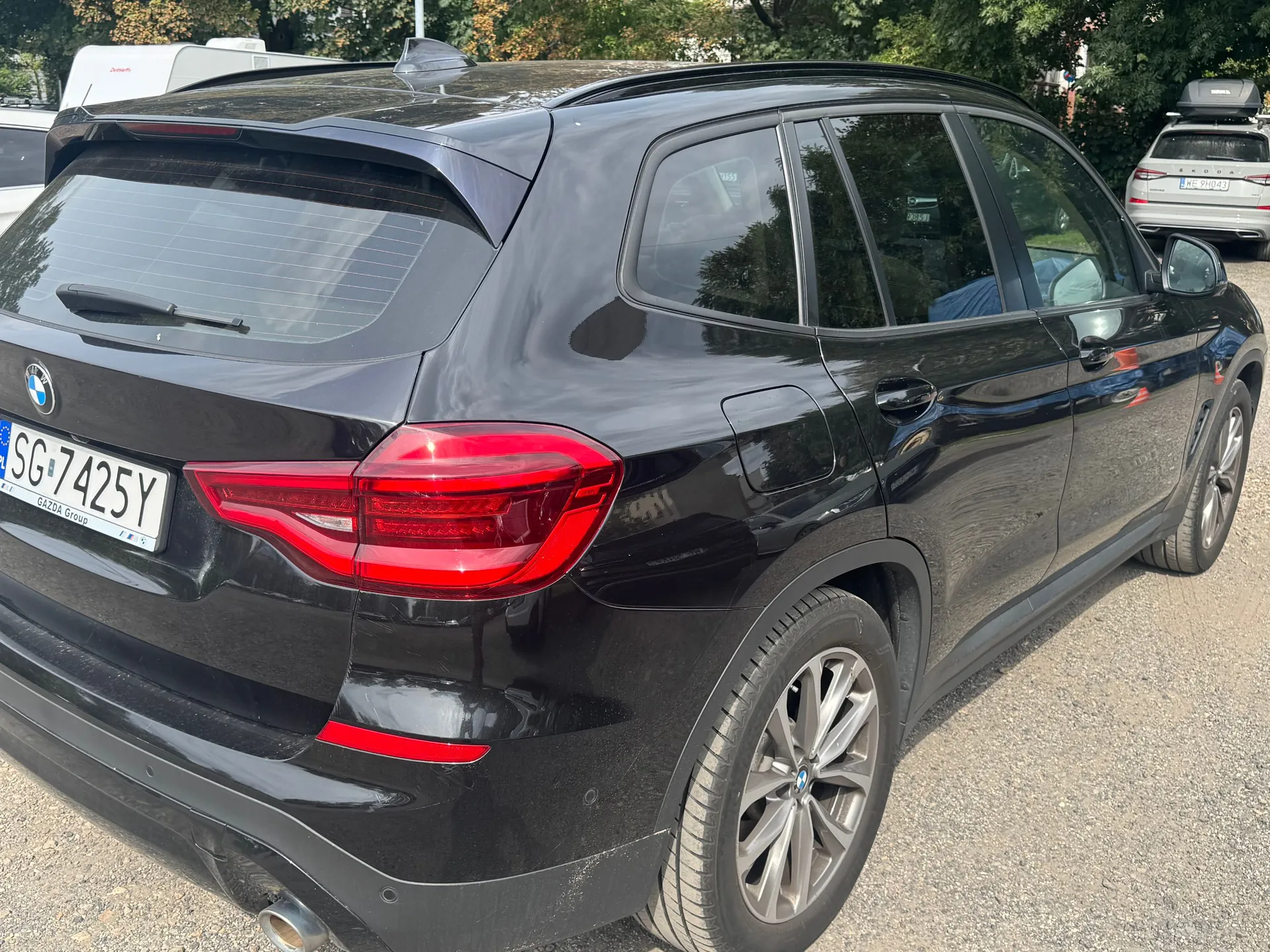 Czarny samochód marki BMW typu SUV zaparkowany na żwirowym placu. Widok z tyłu i z boku pojazdu. Samochód ma polską tablicę rejestracyjną o numerze SG 7425Y. W tle widoczne są inne zaparkowane samochody oraz zielone drzewa.