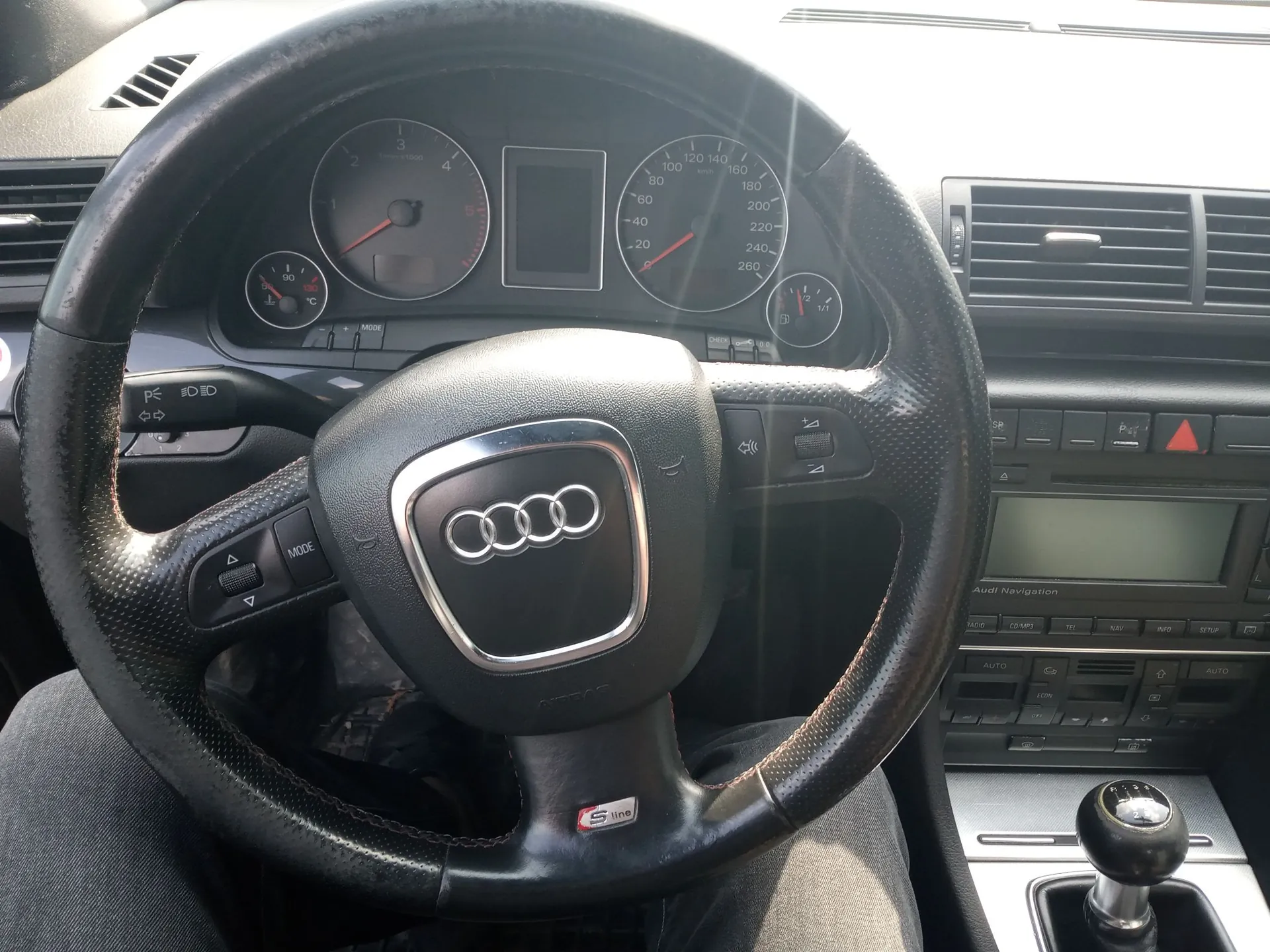 Widok z perspektywy kierowcy na wnętrze samochodu marki Audi. W centrum znajduje się czarna, skórzana kierownica z logo Audi na środku i oznaczeniem 'S line' na dolnym ramieniu. Za kierownicą widoczny jest zestaw wskaźników z prędkościomierzem, obrotomierzem oraz ekranem pokładowym. Po prawej stronie znajduje się konsola środkowa z systemem Audi Navigation, przyciskami sterowania klimatyzacją oraz lewarkiem zmiany biegów.