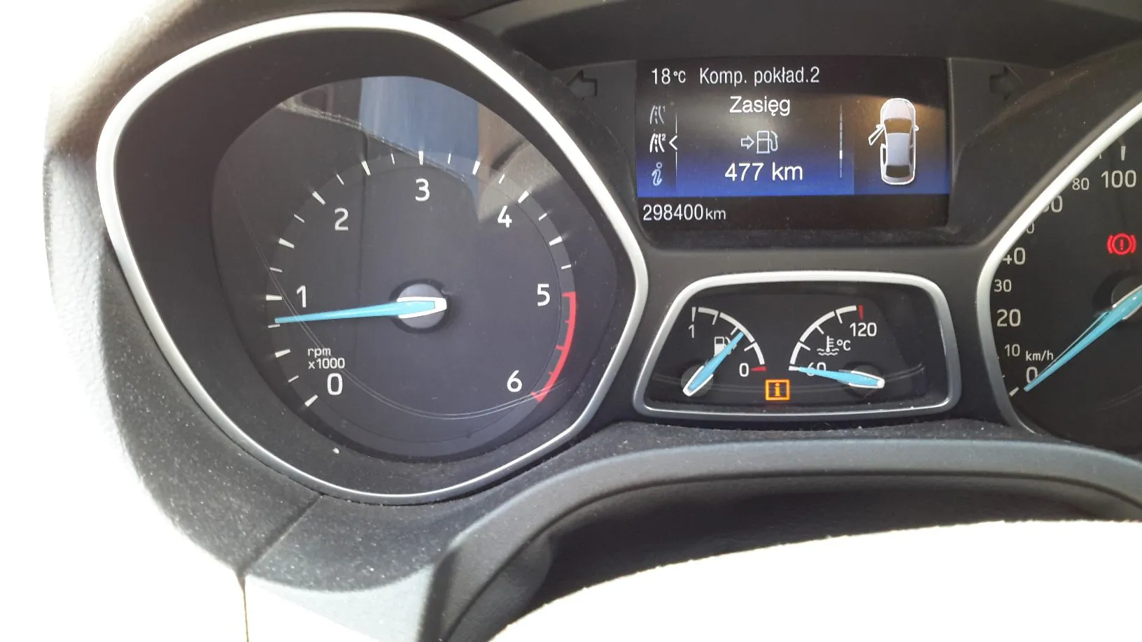 Zbliżenie na deskę rozdzielczą samochodu. Po lewej znajduje się obrotomierz z zaznaczoną skalą od 0 do 6 (rpm x1000). W centralnej części górnej znajduje się ekran komputera pokładowego z tekstem: '18°C Komp. pokład.2', 'Zasięg', '477 km' oraz przebiegiem '298400km' i grafiką samochodu. Poniżej ekranu widoczne są dwa mniejsze wskaźniki: temperatura paliwa i temperatura cieczy chłodzącej (skala do 120°C). Po prawej stronie widoczny jest fragment prędkościomierza z zaznaczoną skalą do 100 km/h.